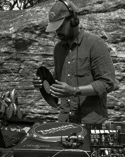 A man wearing headphones and a baseball cap, standing outdoors near a rock wall, appears to be DJing with vinyl records and DJ equipment.