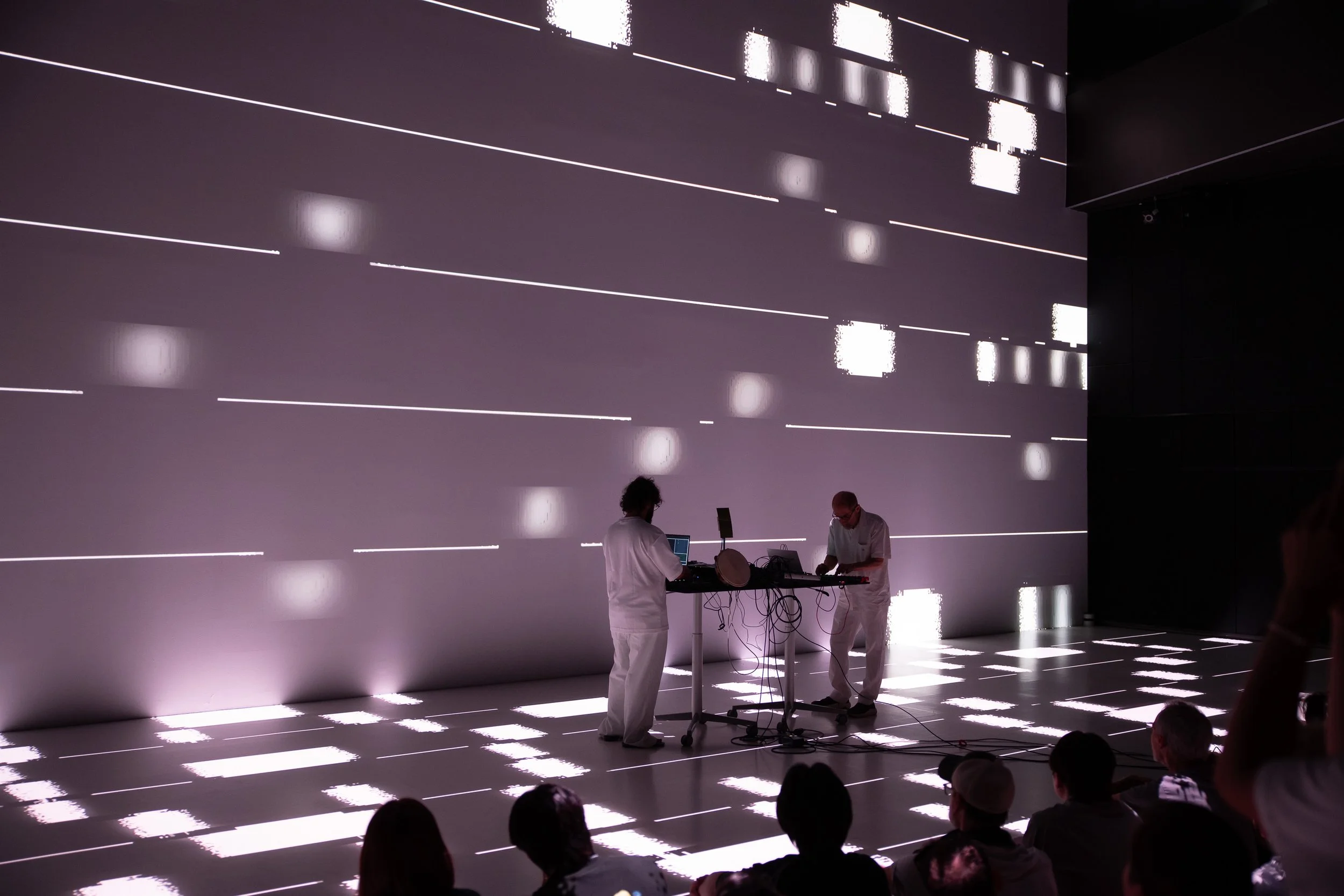 Two performers in white clothing operating electronic music equipment on a stage with white dotted and line lighting effects, while an audience watches in the dark.