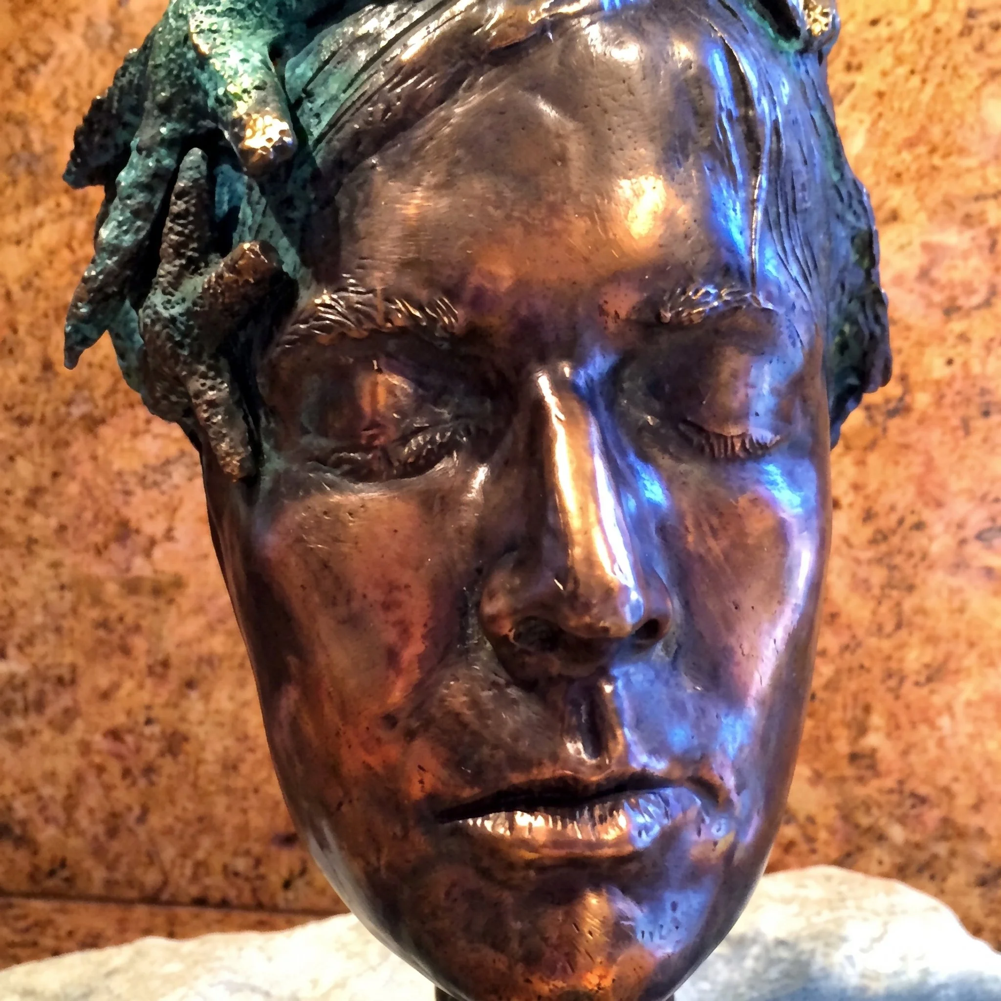 Close-up image of a shiny metallic sculpture of a human face, with detailed features and a reflective surface, set against a blurred background.