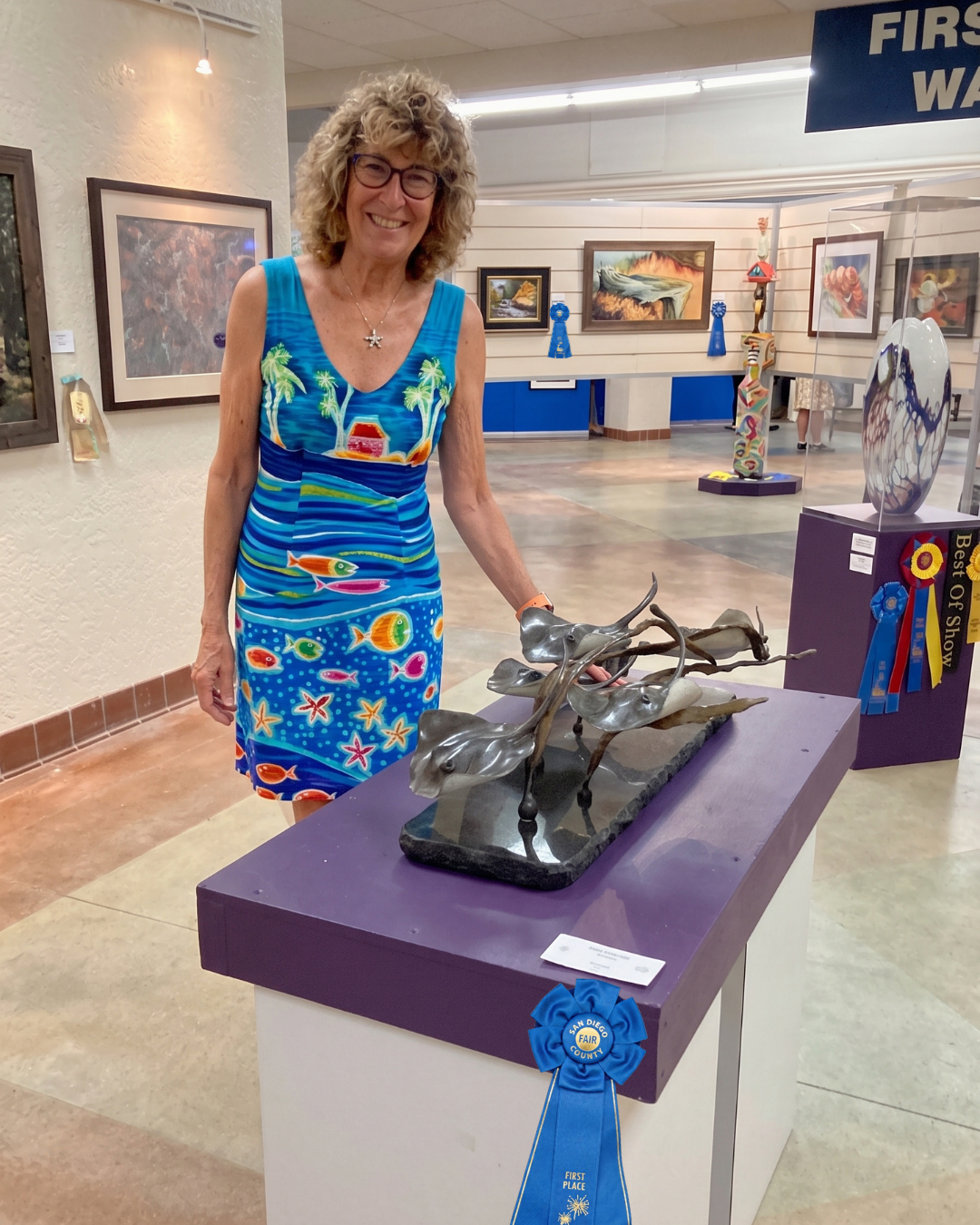 A woman with curly blonde hair and glasses, wearing a colorful dress with ocean and beach themes, stands next to a sculpture of three metal frogs on a purple display table that has a blue first place ribbon. The art exhibit features paintings and sculptures in the background, with a sign reading 'First' visible.
