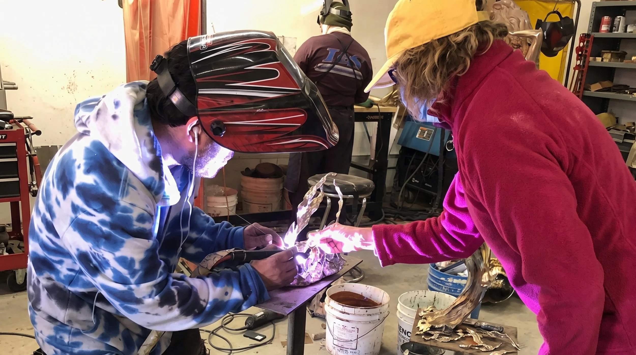 Judy and friend working on a sculpture, one wearing a welding helmet and the other wearing a yellow hat, in a foundry workshop.
