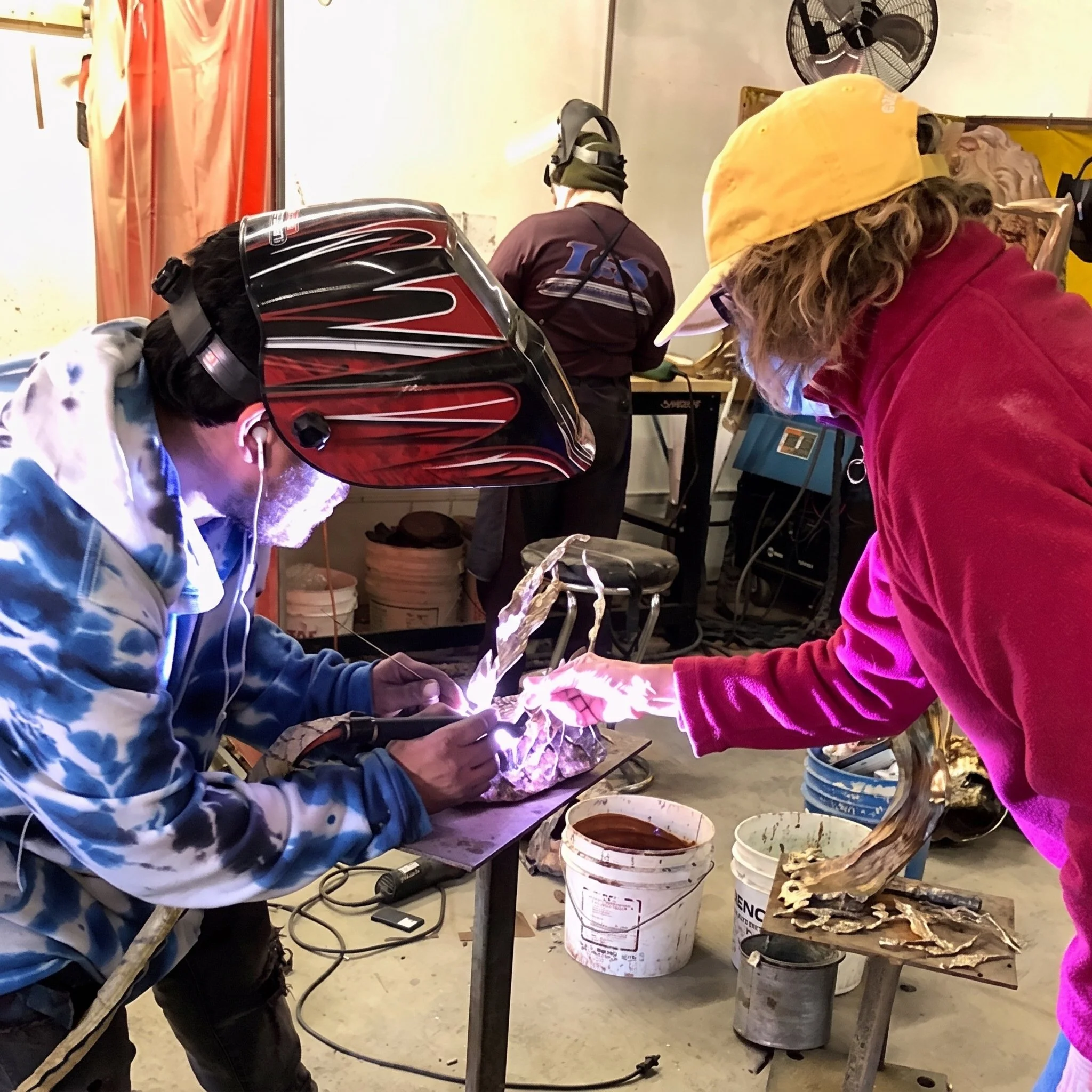 Individual wearing a motorcycle helmet and gloves working on a project using a welding machine in a workshop, with various buckets and materials around.
