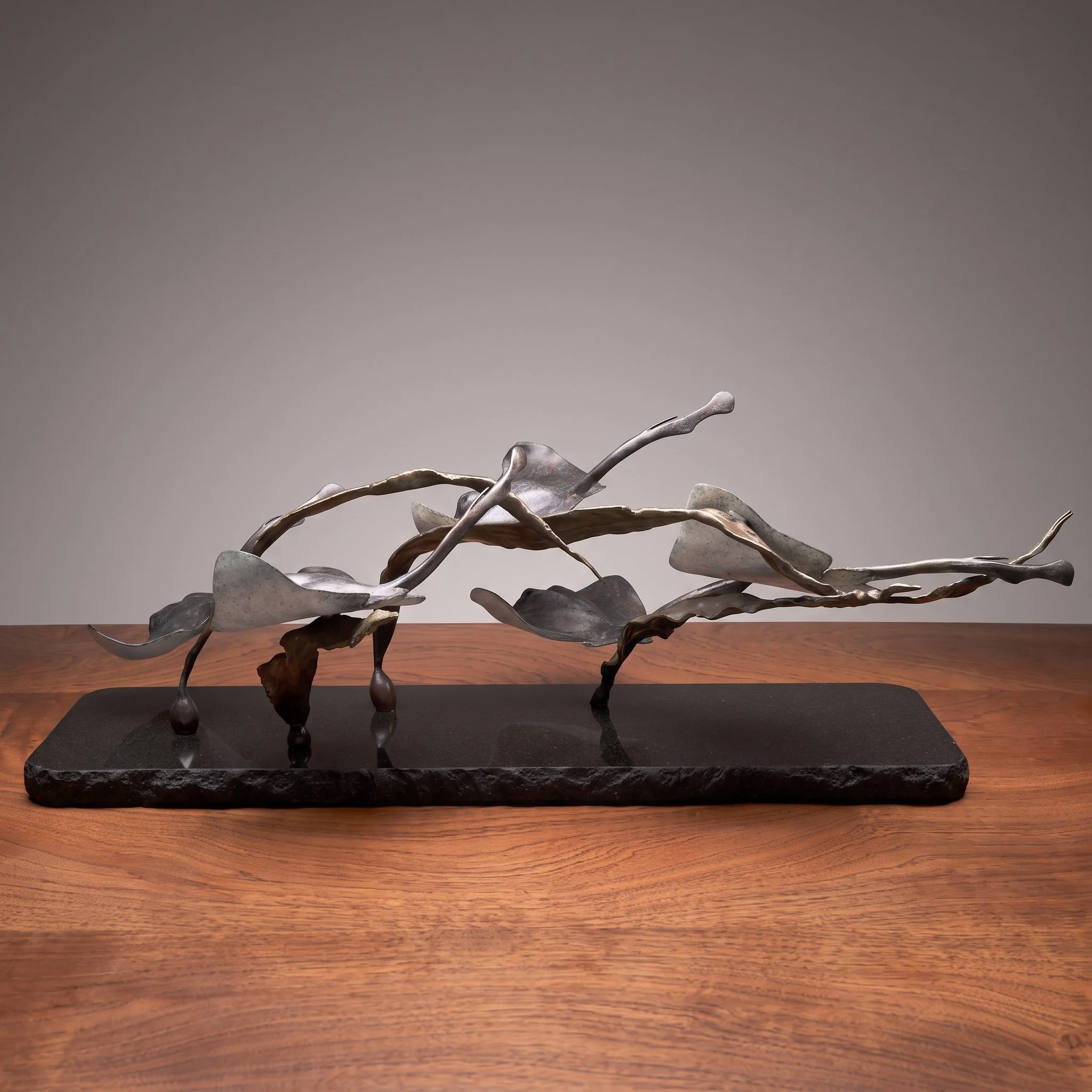 A metallic sculpture resembling twisted and intertwined branches or vines placed on a wooden table against a plain gray background.