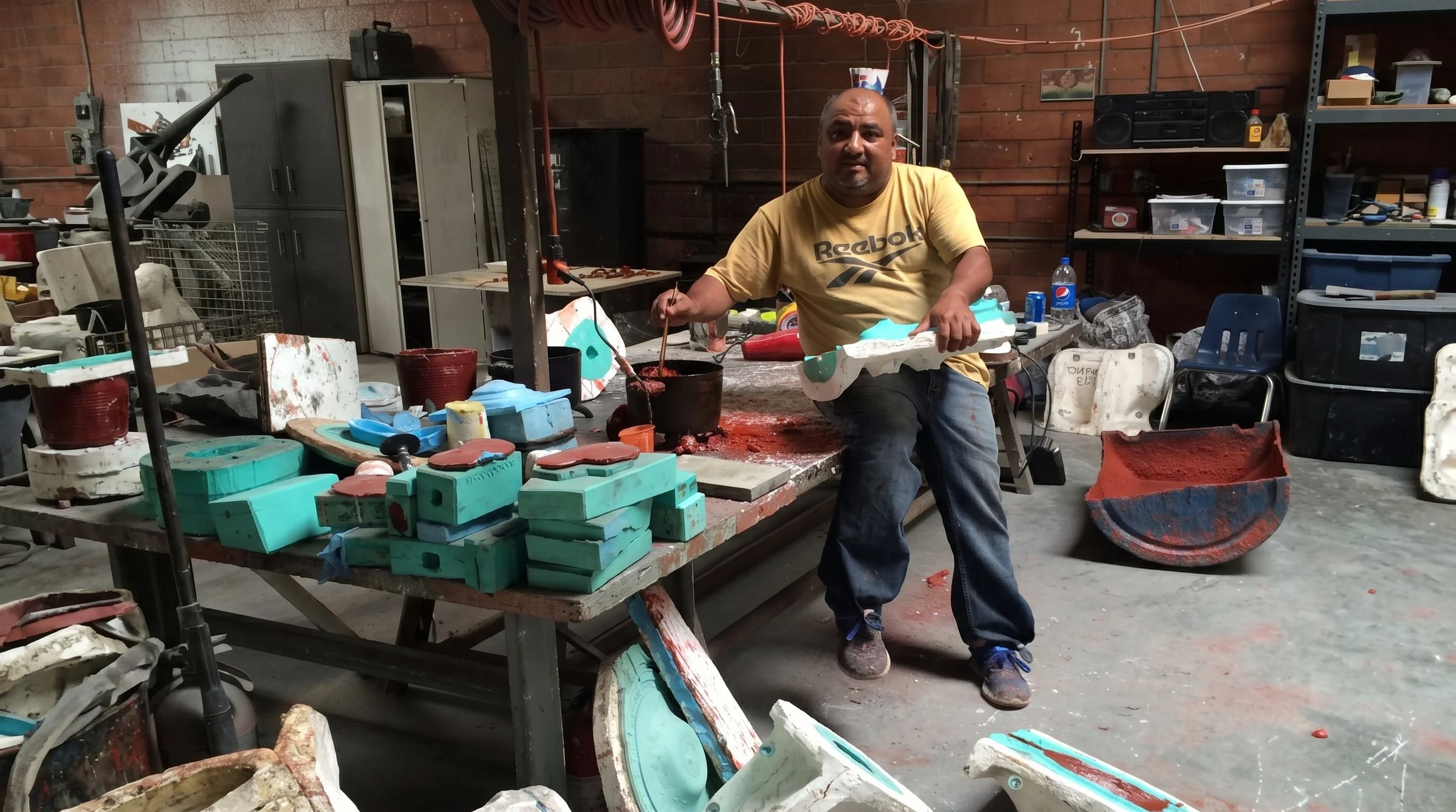 A man in a yellow Reebok shirt working in a cluttered workshop with foam molds and tools on a table, brick walls, shelves with boxes, and various equipment in the background.