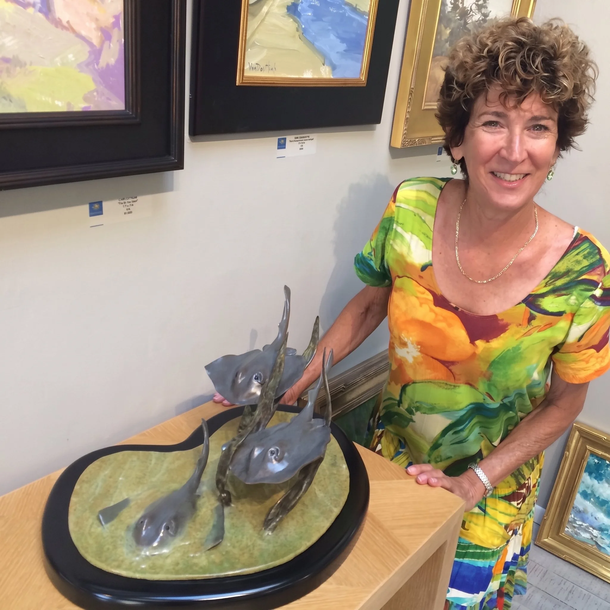 A woman with curly hair and a colorful dress smiling at an art gallery, standing next to a metallic sculpture of deer antlers on a green surface.