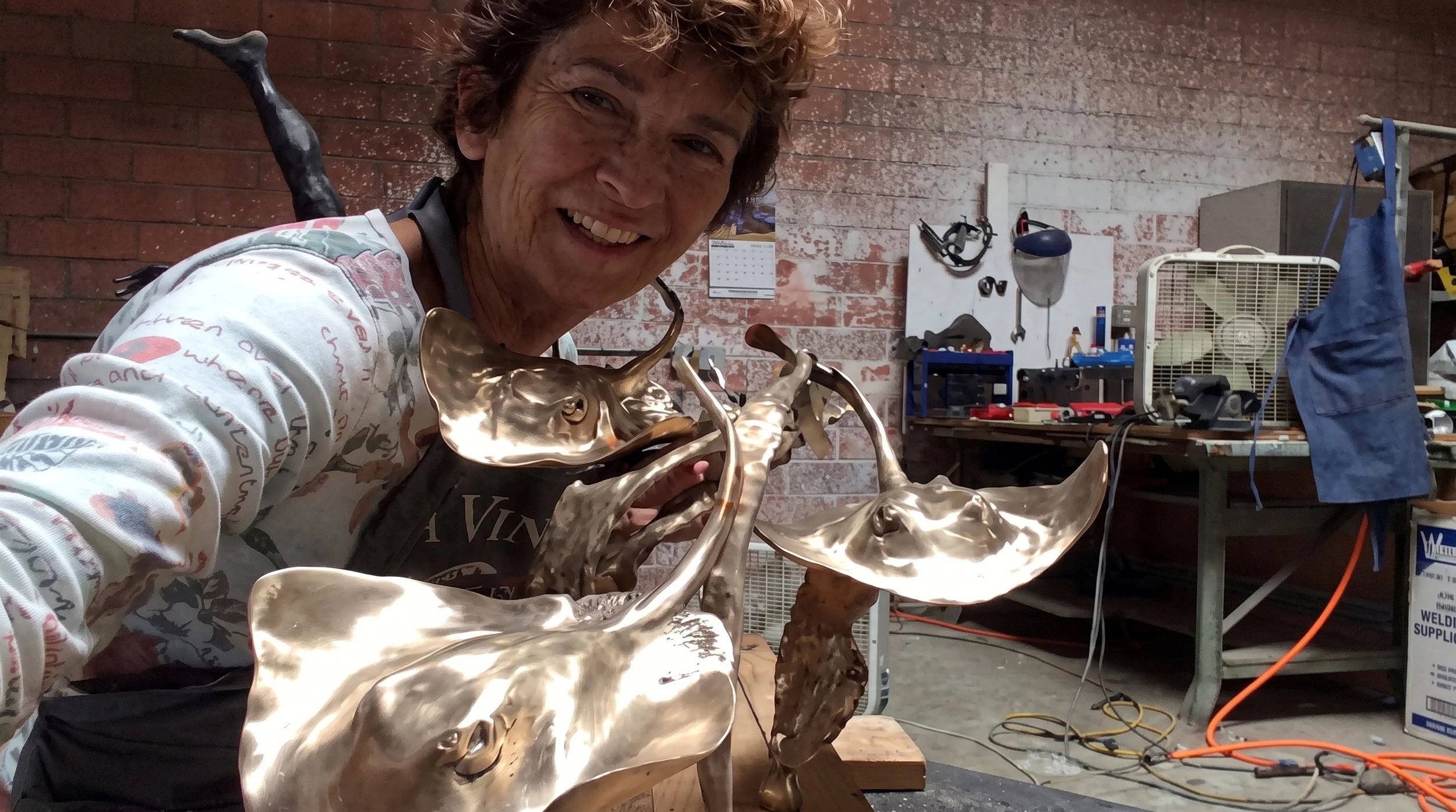 Judy Salinsky smiling at the camera while holding a large, shiny, stingray sculpture in a workshop with brick walls, tools, and equipment in the background.