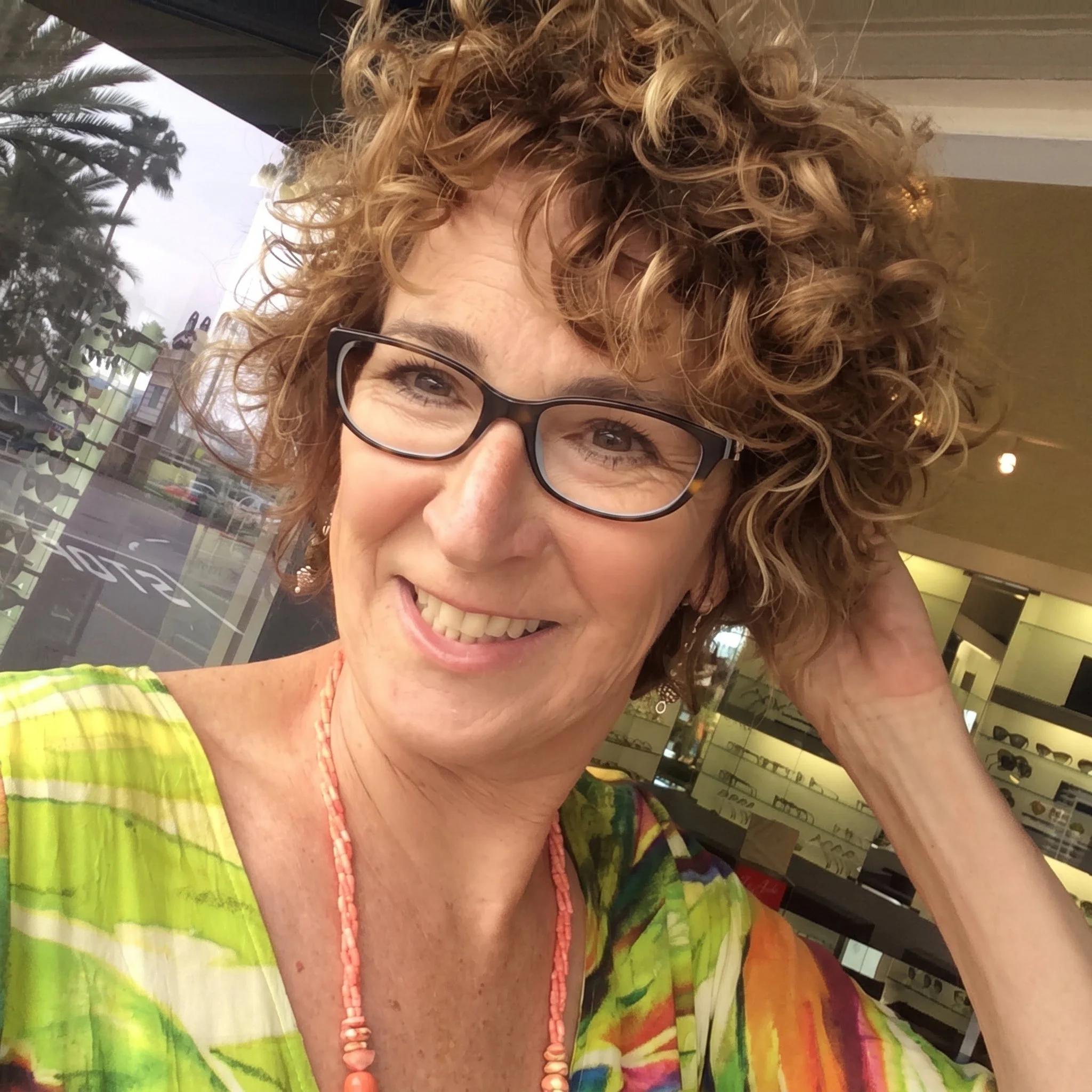 A woman with curly red hair and glasses, smiling inside a store, wearing a colorful tie-dye shirt and a coral beaded necklace.