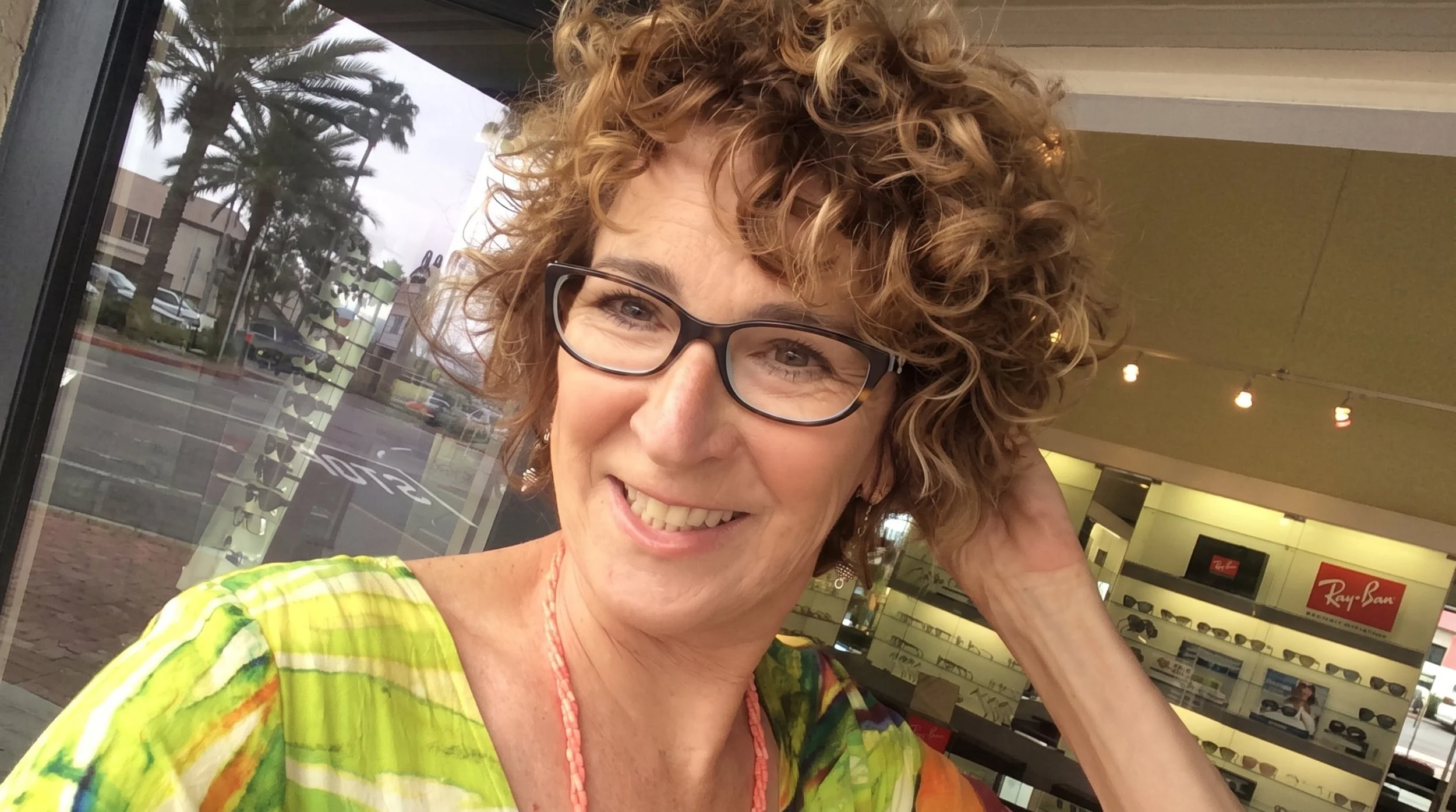 A woman with curly blonde hair and glasses smiling inside a store with sunglasses display and Big Palm trees outside the window.