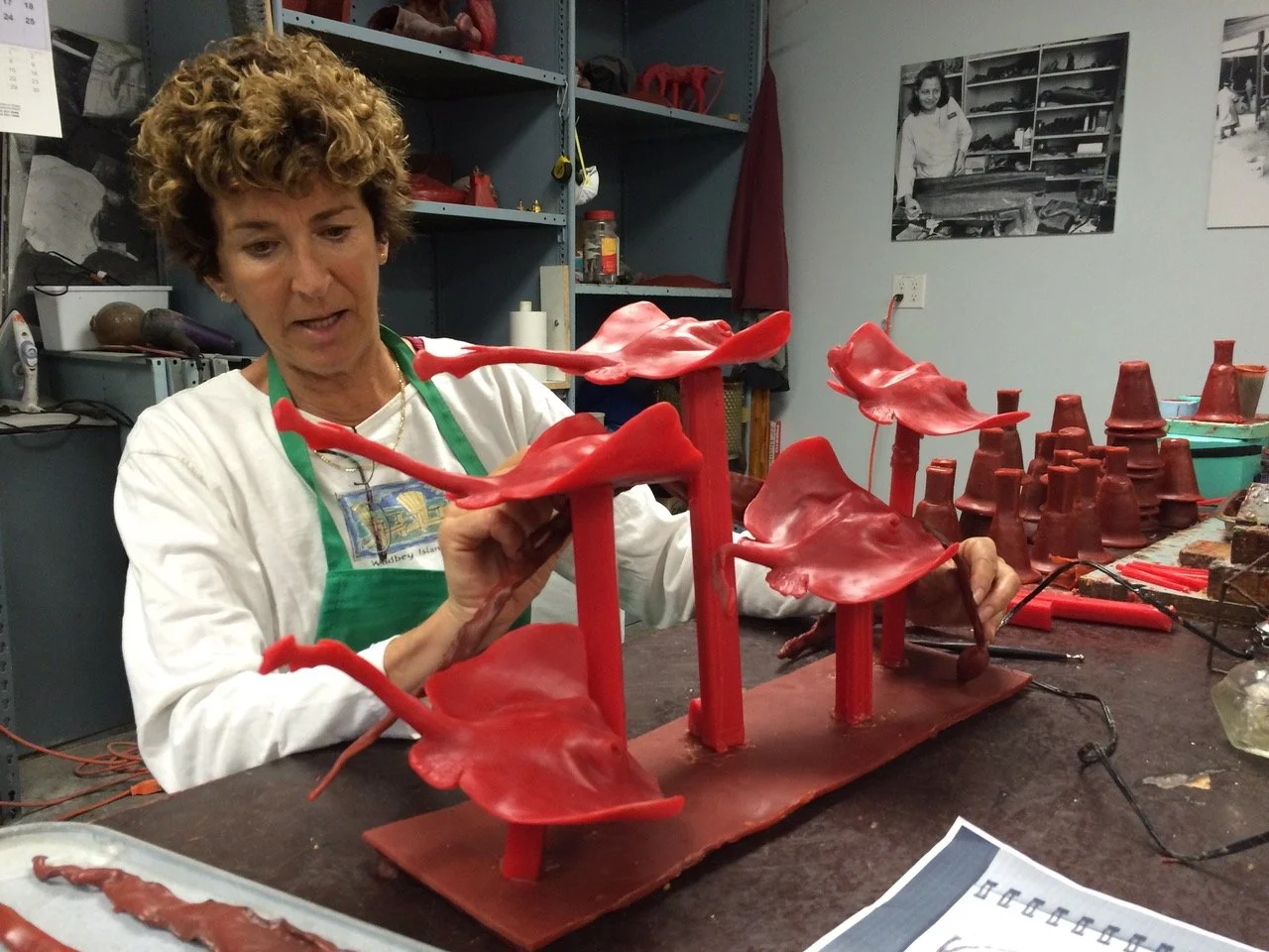 Judy Salinsky working on a red stingray sculpture resembling large flower petals on a workshop table, with shelves and photographs in the background.