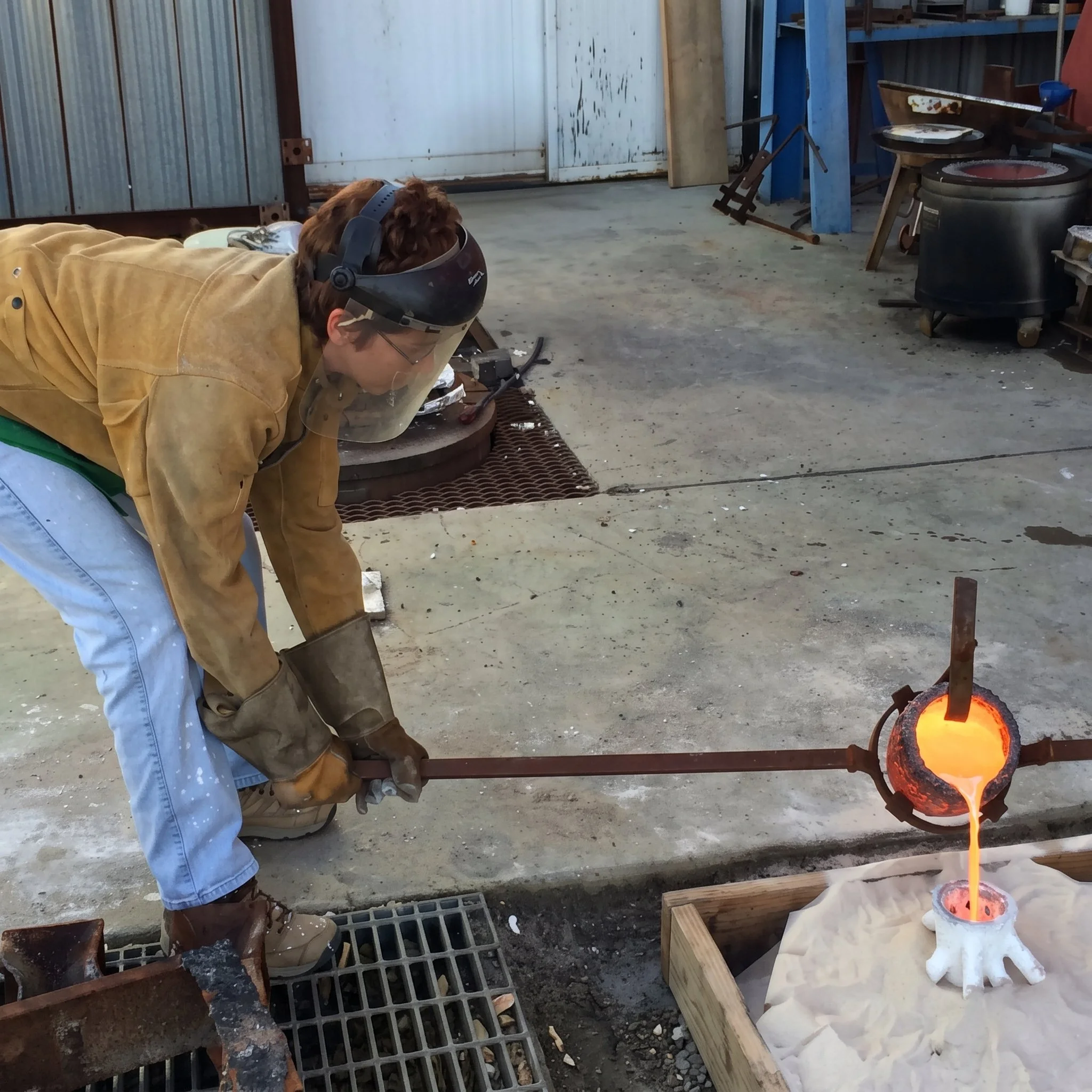 Judy Salinsky the artist pours hot bronze liquid into mold for a project she's working on, in the Foundry of Berkeley , wearing safety goggles and a yellow jacket, with glowing molten glass on a table and various tools and equipment around.