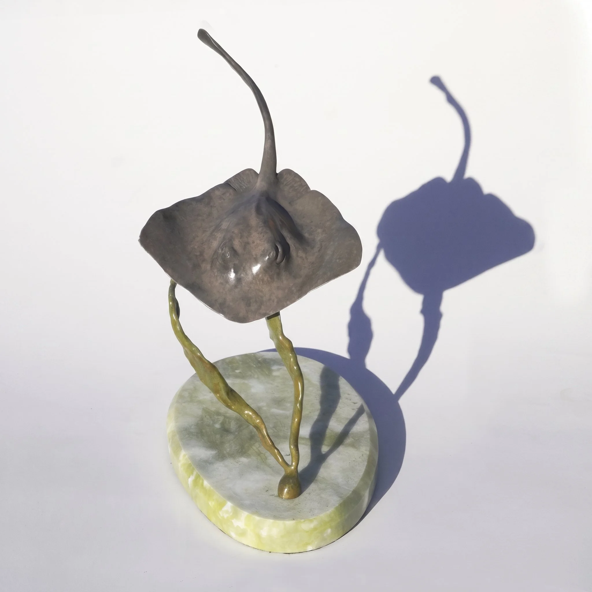 A bronze sculpture of a single stingray on a circular stone base, casting a shadow on a white background, hence the name "Shadow Dancer" by Judy Salinsky