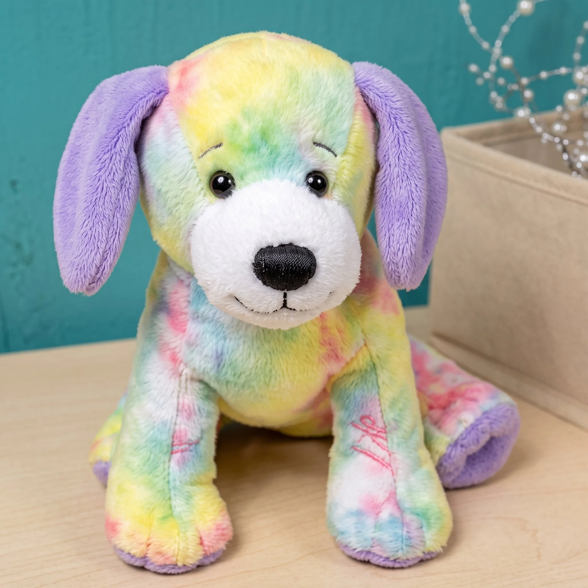 A plush stuffed animal dog with rainbow colors, purple ears, and a white snout, sitting on a wooden surface with a teal background.