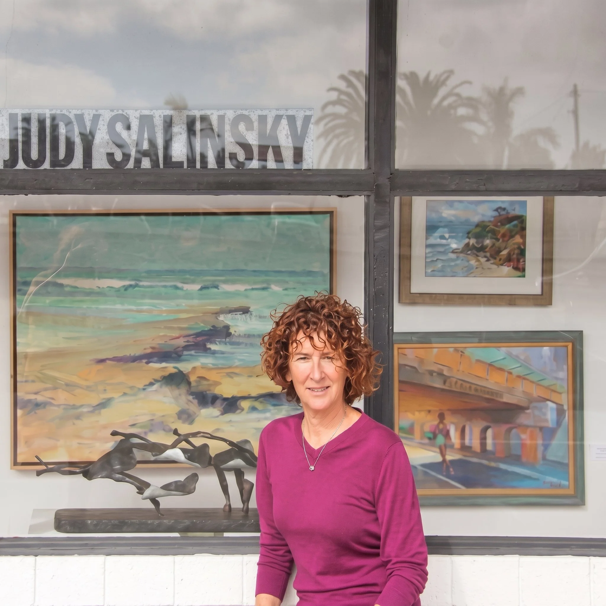 Woman with curly red hair wearing a magenta long-sleeve top standing in front of framed artwork and a sculpture in an art gallery.