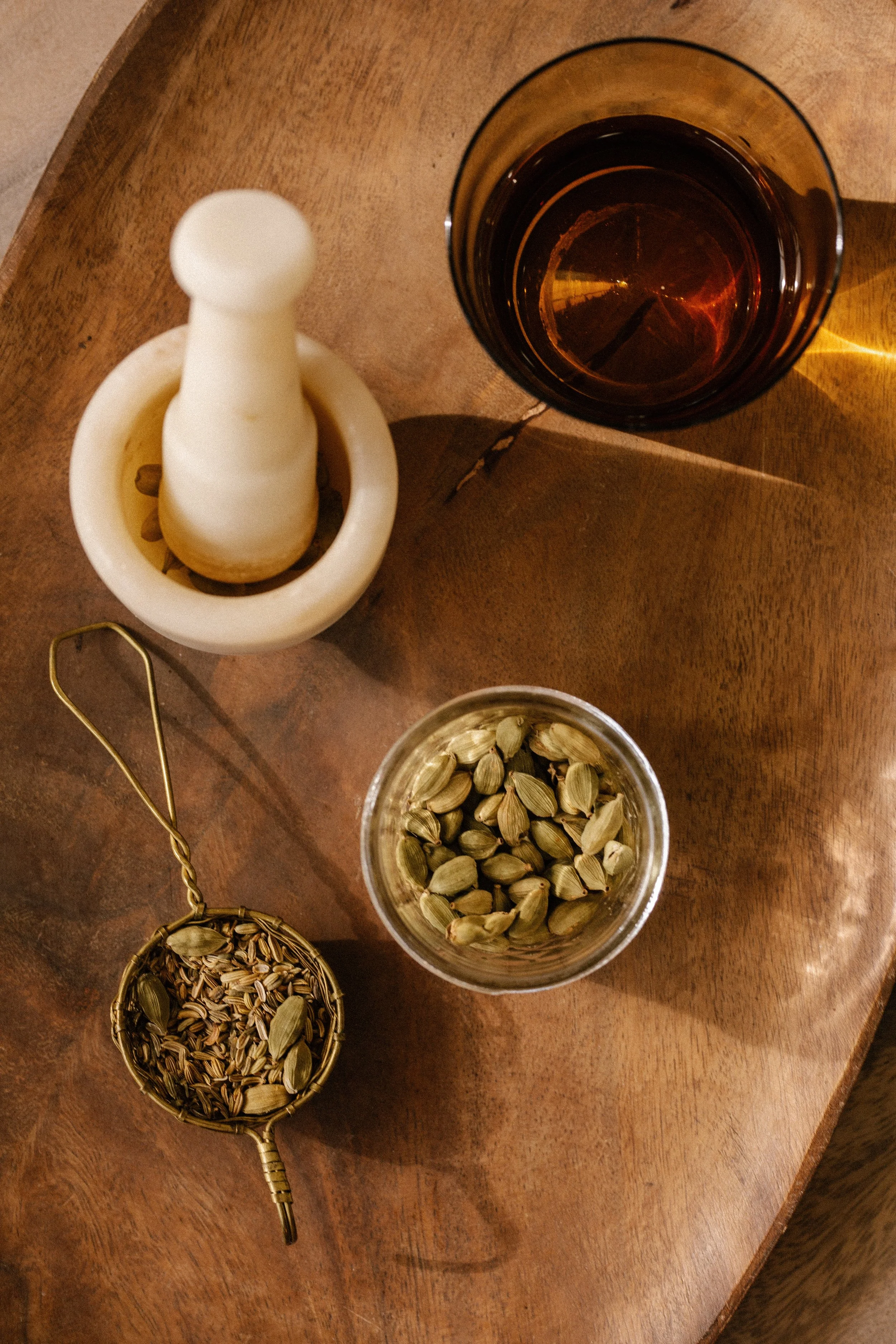 A mortar and pestle, cardamom pods and a tea strainer of seeds on a wooden board