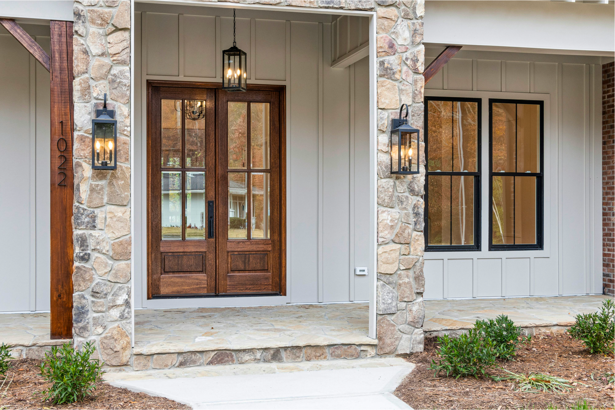 Front porch of a house with a stone and siding exterior, featuring a wooden front door with glass panels, two wall-mounted lantern light fixtures, a small decorative porch, and some green plants in the front yard.