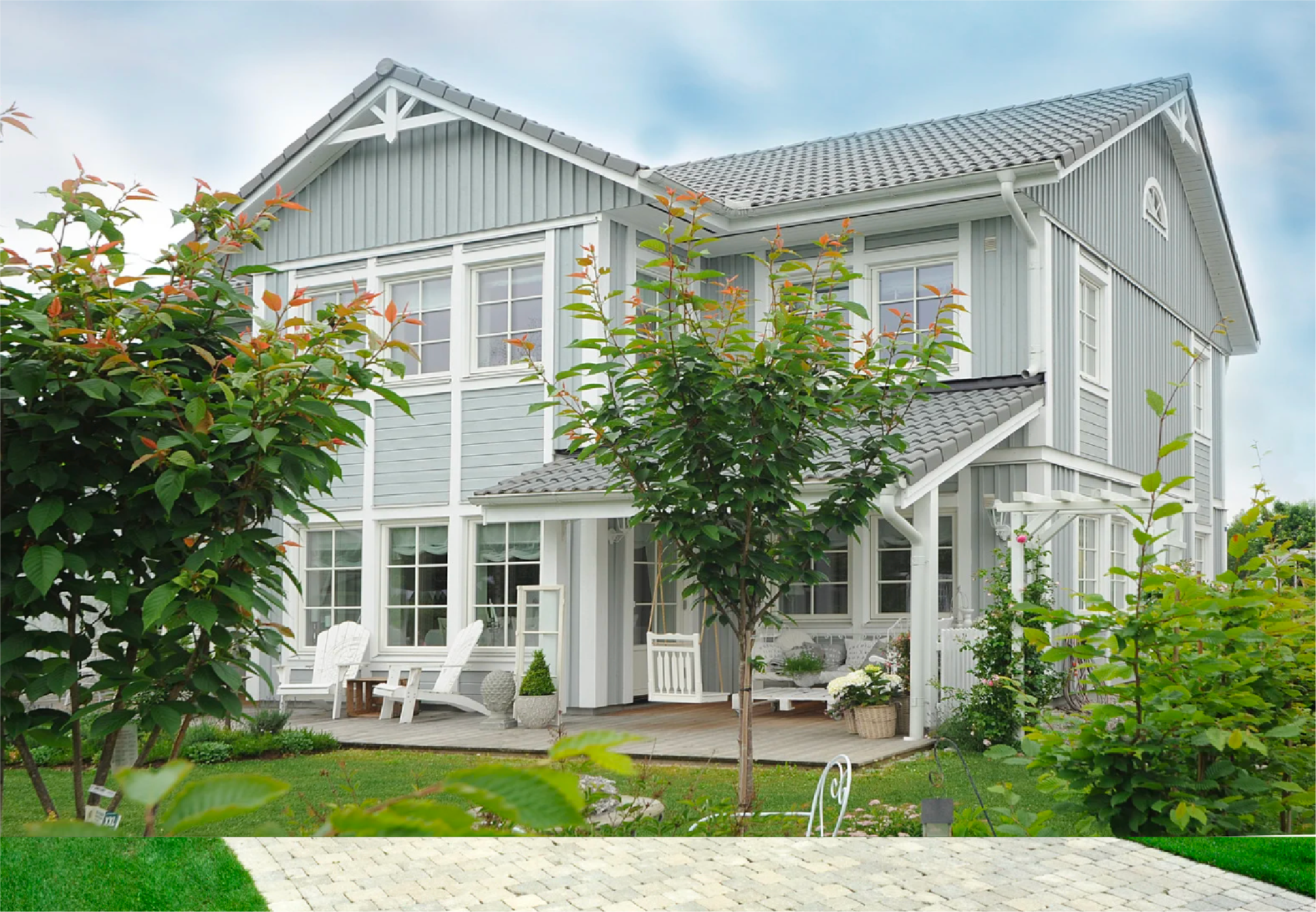 White house with a gray tiled roof, surrounded by greenery including trees, bushes, and a lawn, with a stone pathway leading to the house.