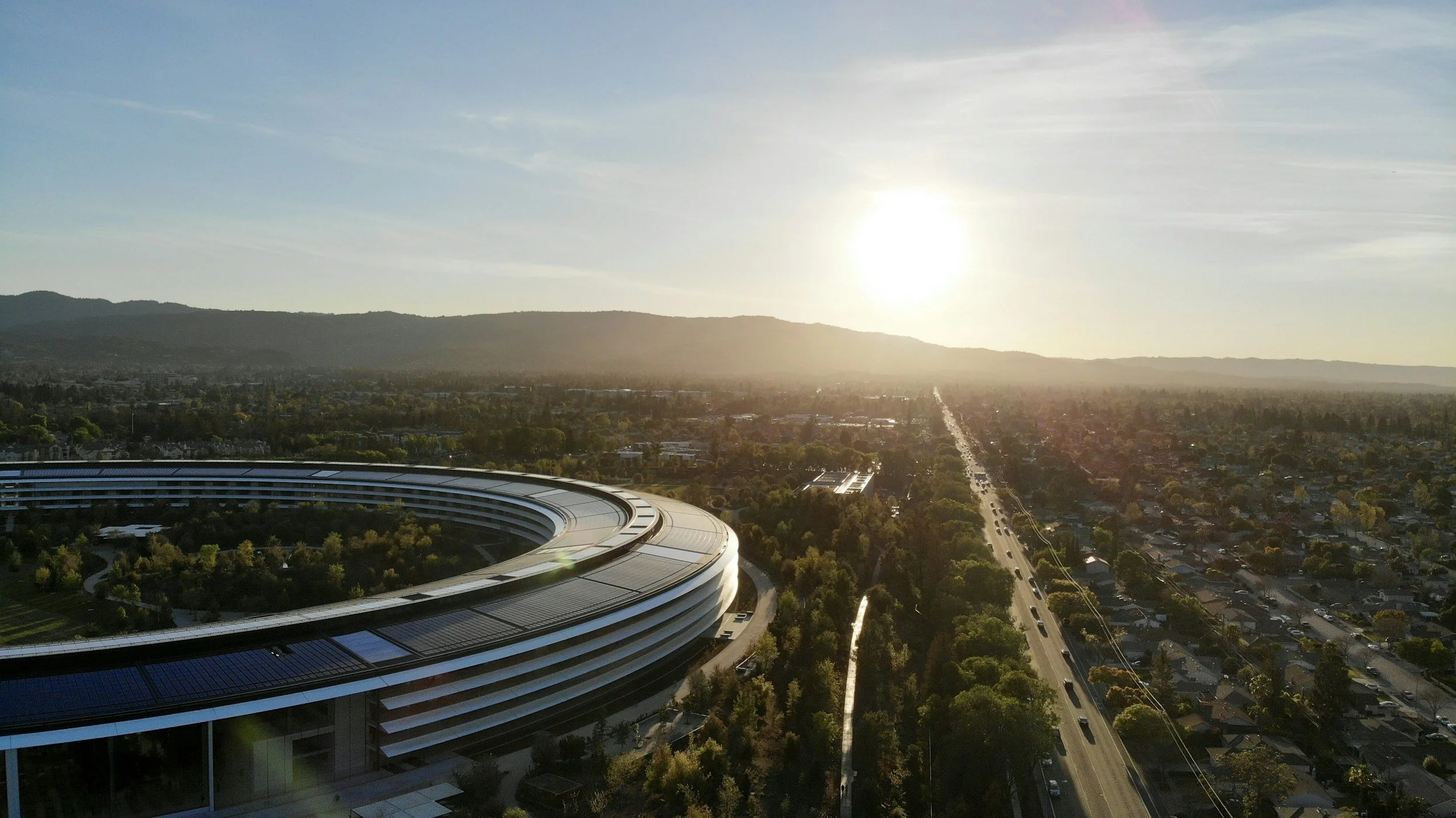 An aerial view of a city landscape during sunset with a futuristic, circular building in the foreground and mountains in the background.