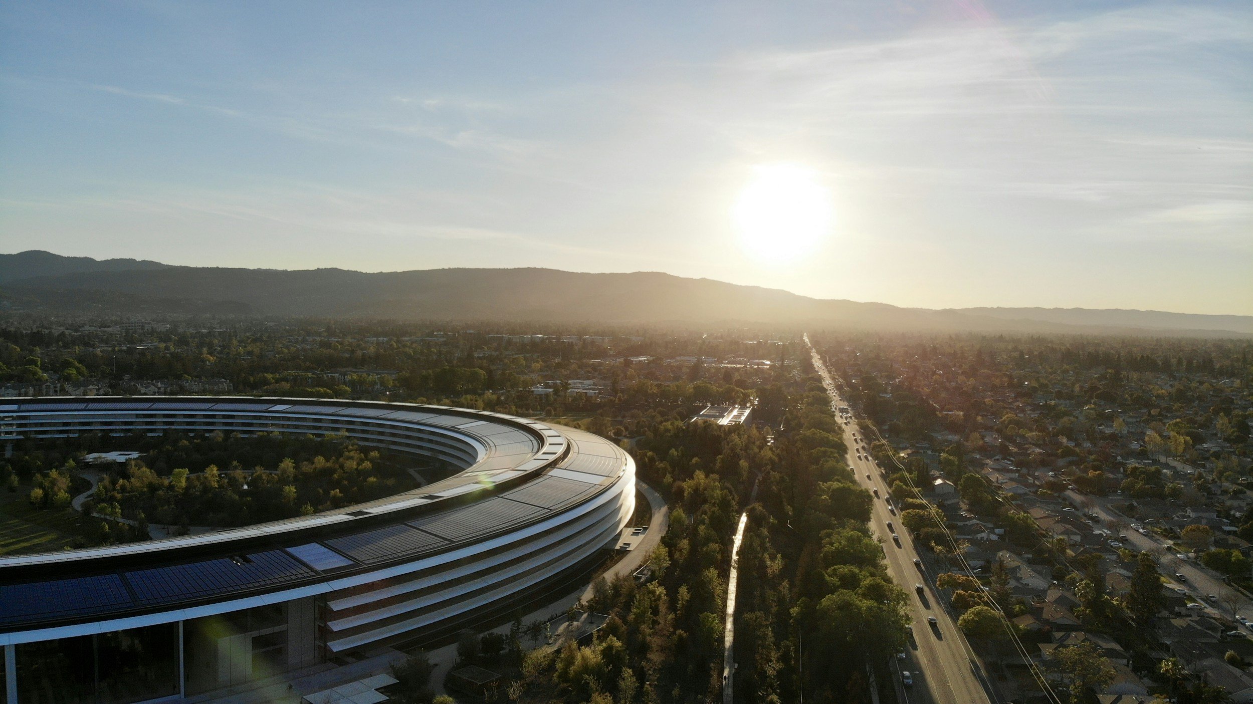 Aerial view of a city with a modern circular building in the foreground and a mountain range in the background, with the sun low in the sky.