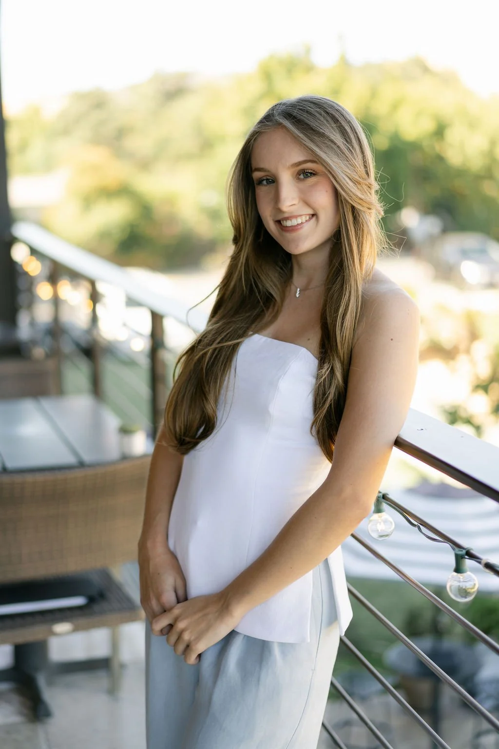 A young woman with long, wavy brown hair smiling and looking at the camera on a balcony during daytime. She is wearing a white sleeveless top and light-colored pants, with a blurred background of trees and outdoor scenery.