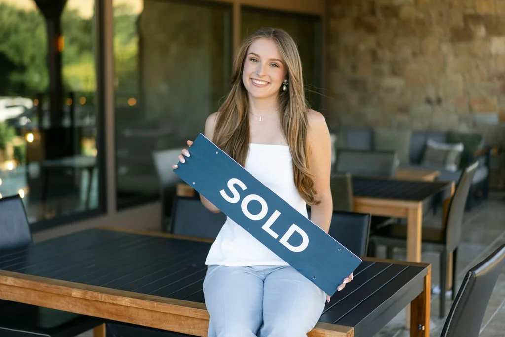 Young woman sitting at a table holding a sign that says 'SOLD' after buying or selling a house.