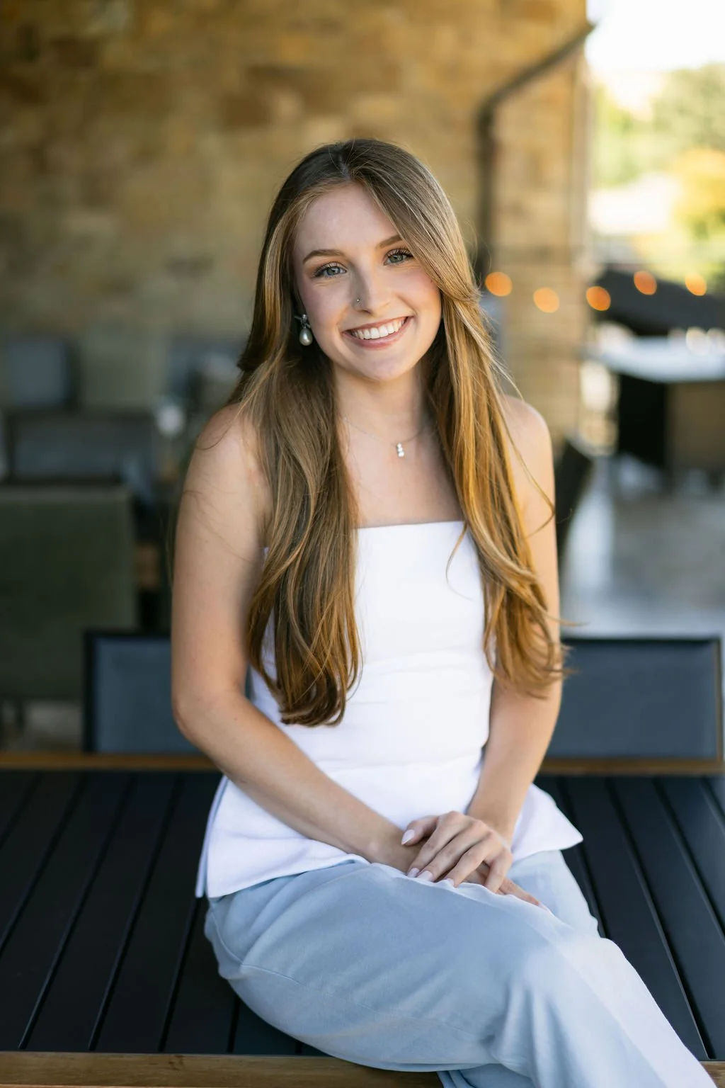 A young woman with long, wavy brown hair, smiling, sitting outdoors on a black wooden bench, wearing a white sleeveless top and light gray pants, with a blurred background of a brick wall and string lights.