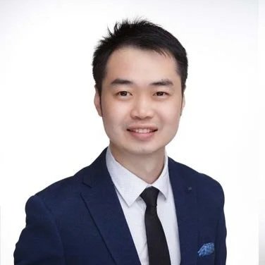 A young man in a navy blue suit, white shirt, and black tie smiling against a plain white background.