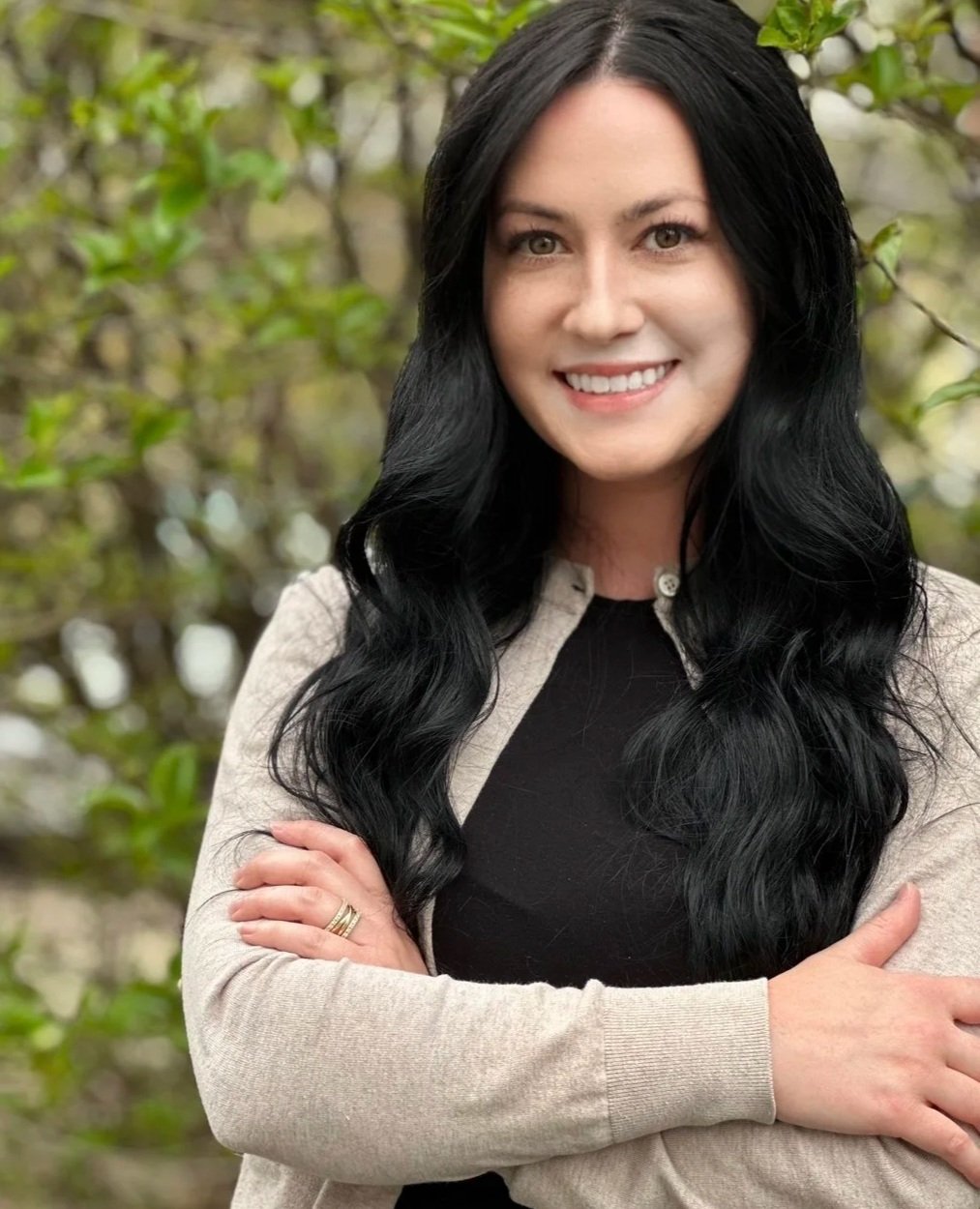 A woman with long black hair, smiling, with her arms crossed, wearing a beige cardigan over a black top, standing outdoors in a natural setting with green foliage in the background.