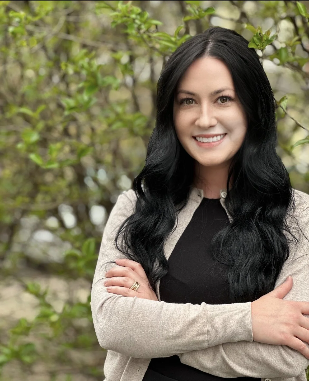 A woman with long black hair smiling outdoors, standing in front of green leafy bushes.