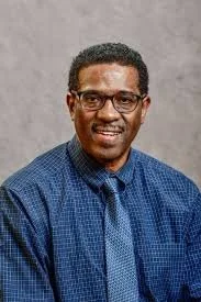 A man with glasses and short curly hair wearing a blue button-up shirt, smiling against a neutral gray background.