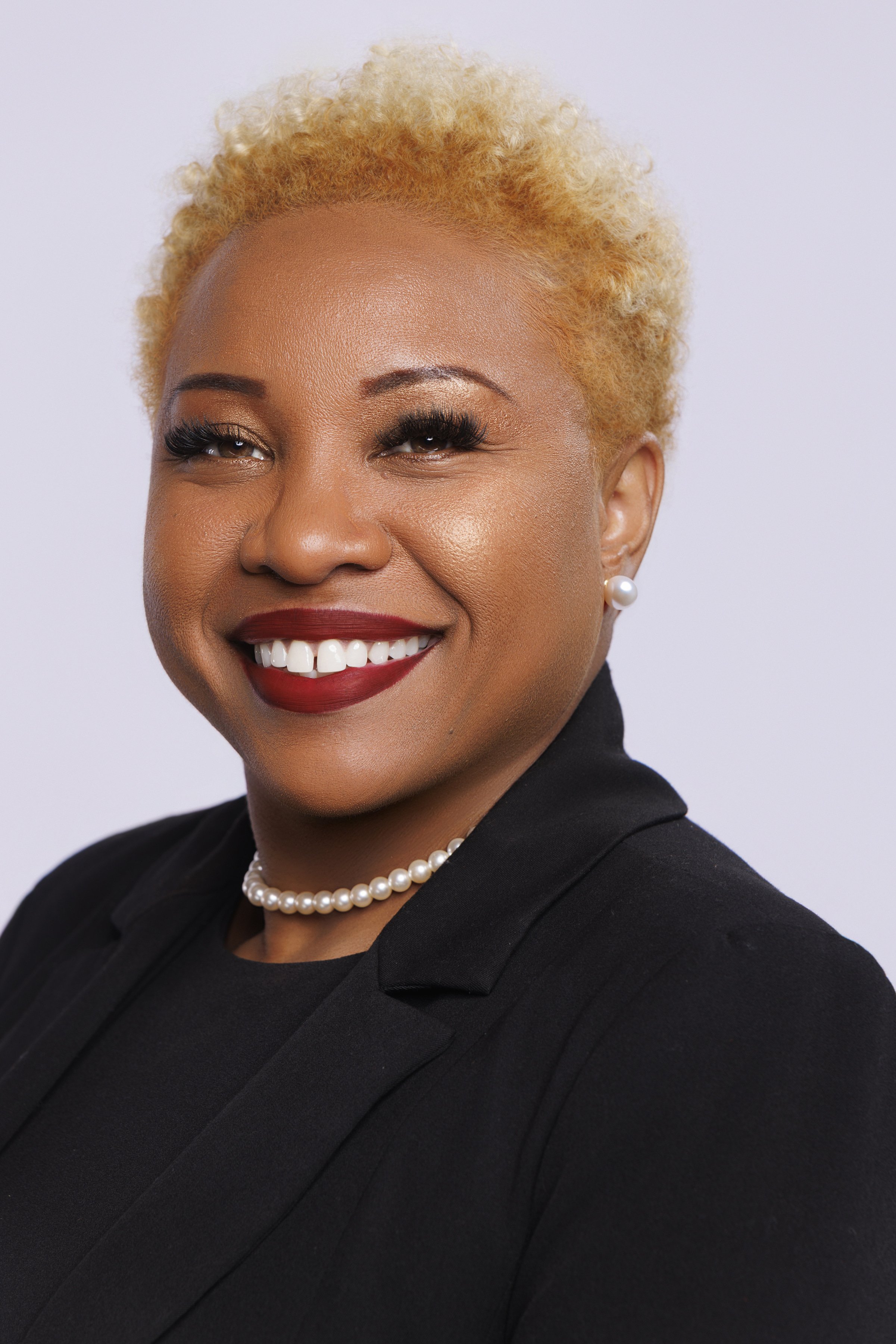 A woman with short, styled blonde hair, wearing a black blazer, pearl necklace, pearl earrings, and red lipstick, smiling against a plain light background.
