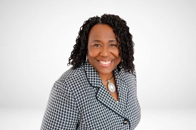 A smiling African American woman with curly hair wearing a black and white patterned blazer and a large silver necklace, standing against a plain white background.