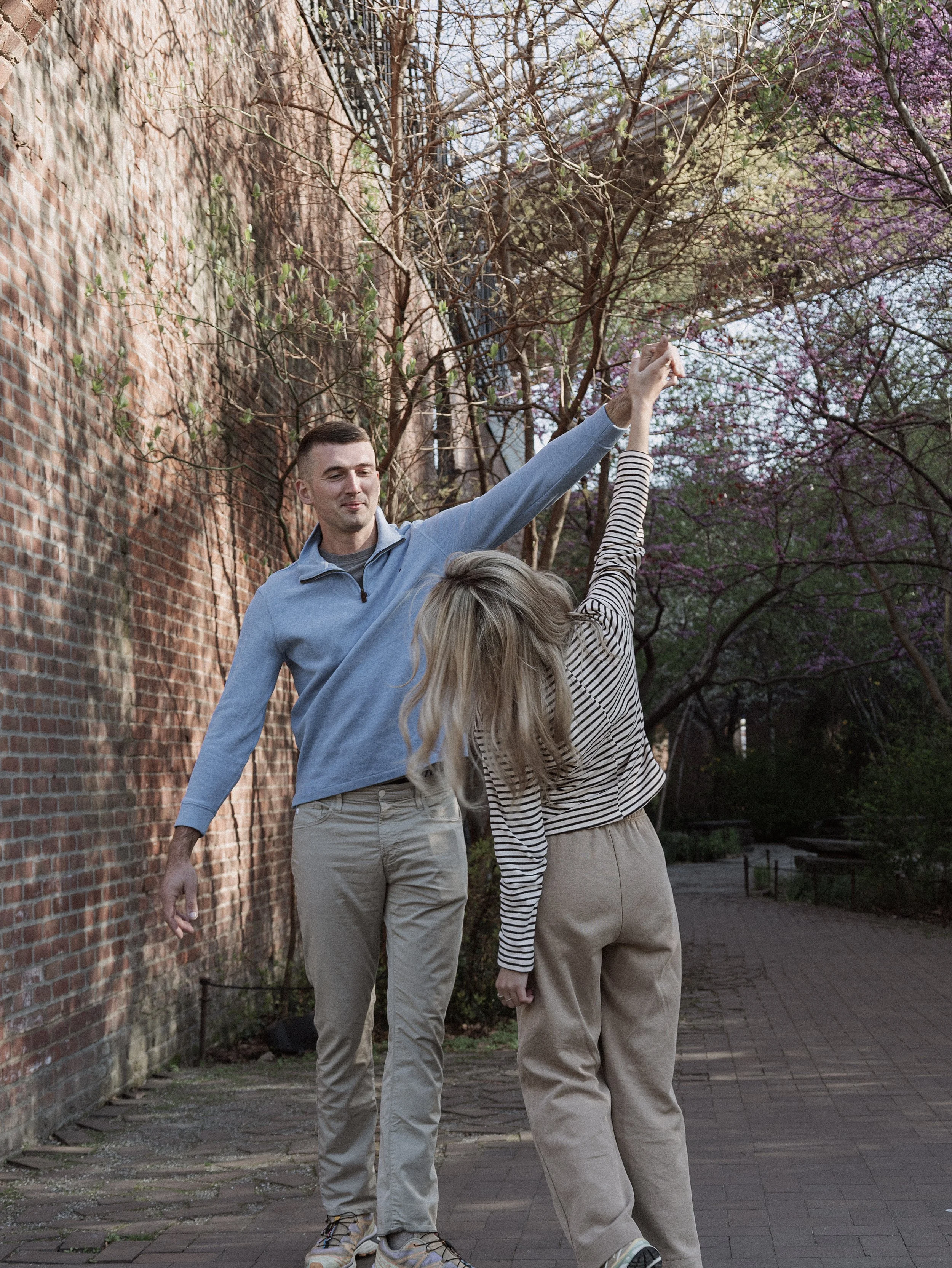 Two people with raised arms and intertwined hands standing outdoors near a brick wall and colorful blooming trees.