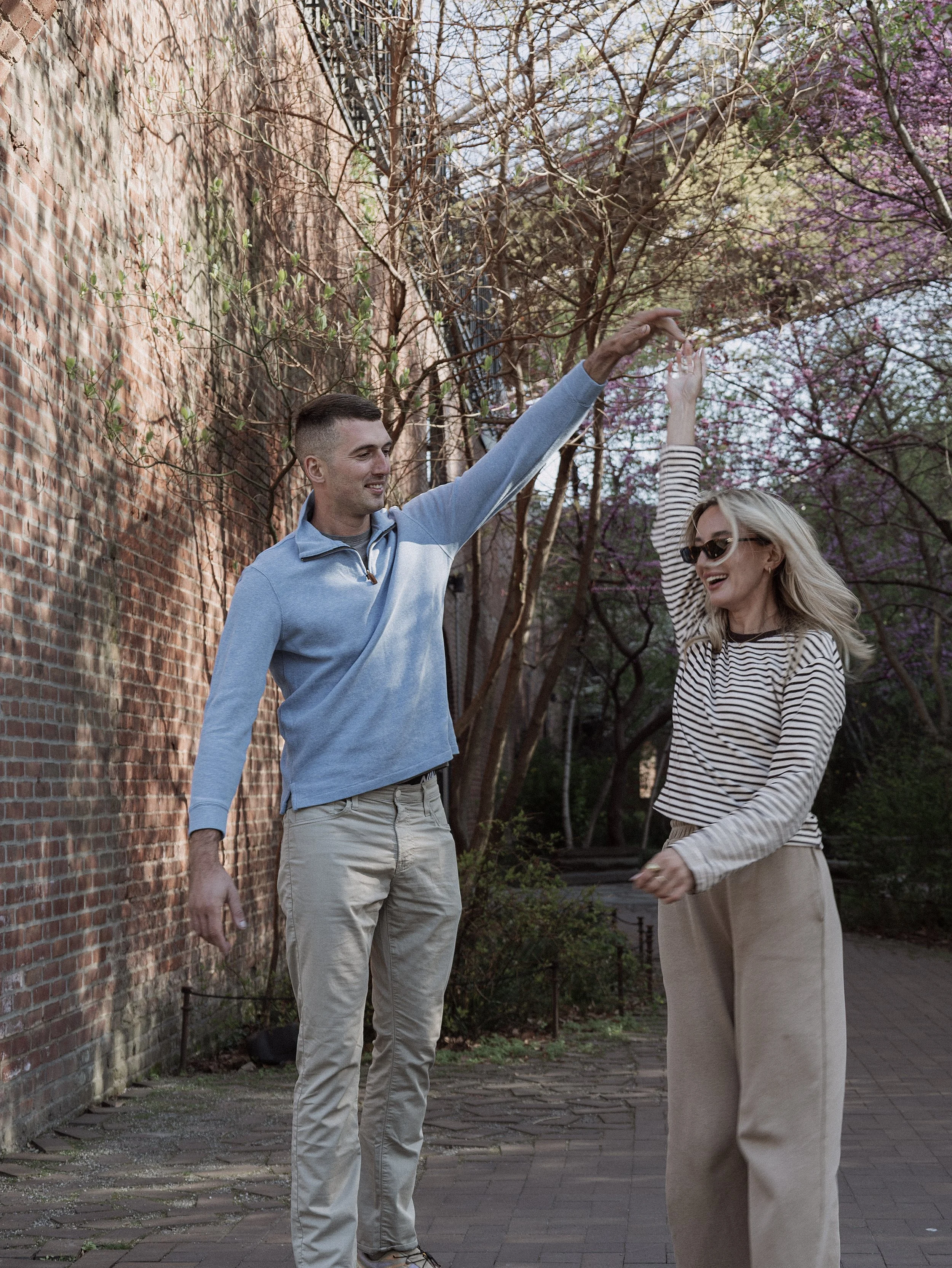 A man and a woman are outdoors near a brick wall and trees, dancing or playing together, with the man raising his hand and the woman smiling.