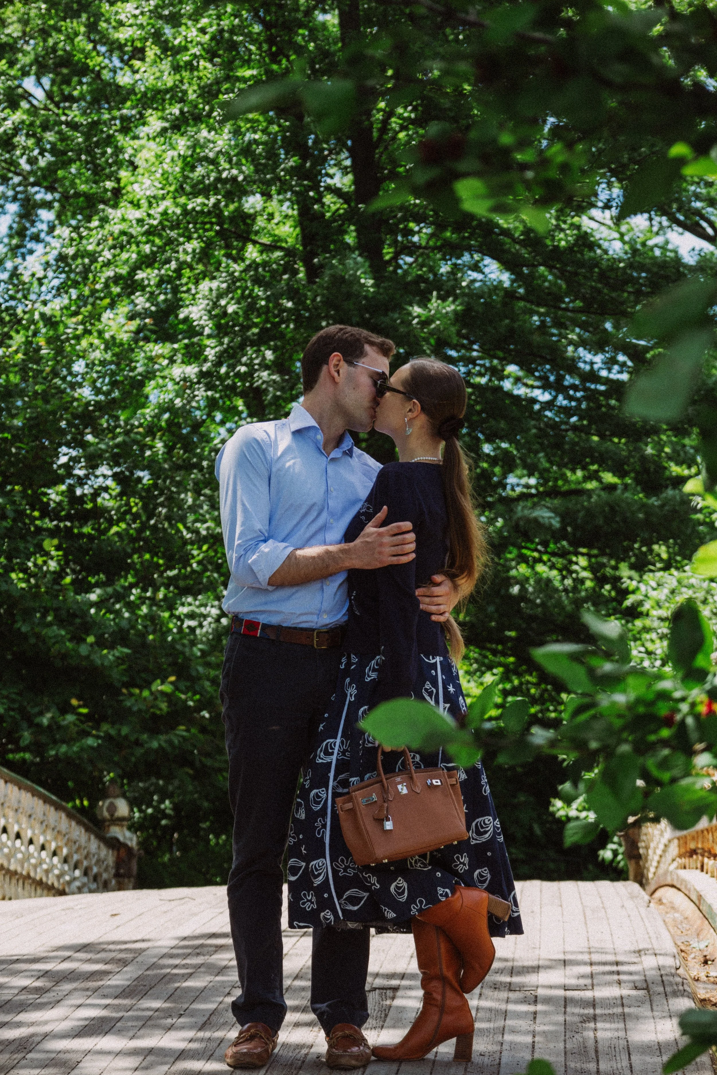 A couple kissing outdoors surrounded by green trees, with a wicker fence and a wooden walkway in the background.