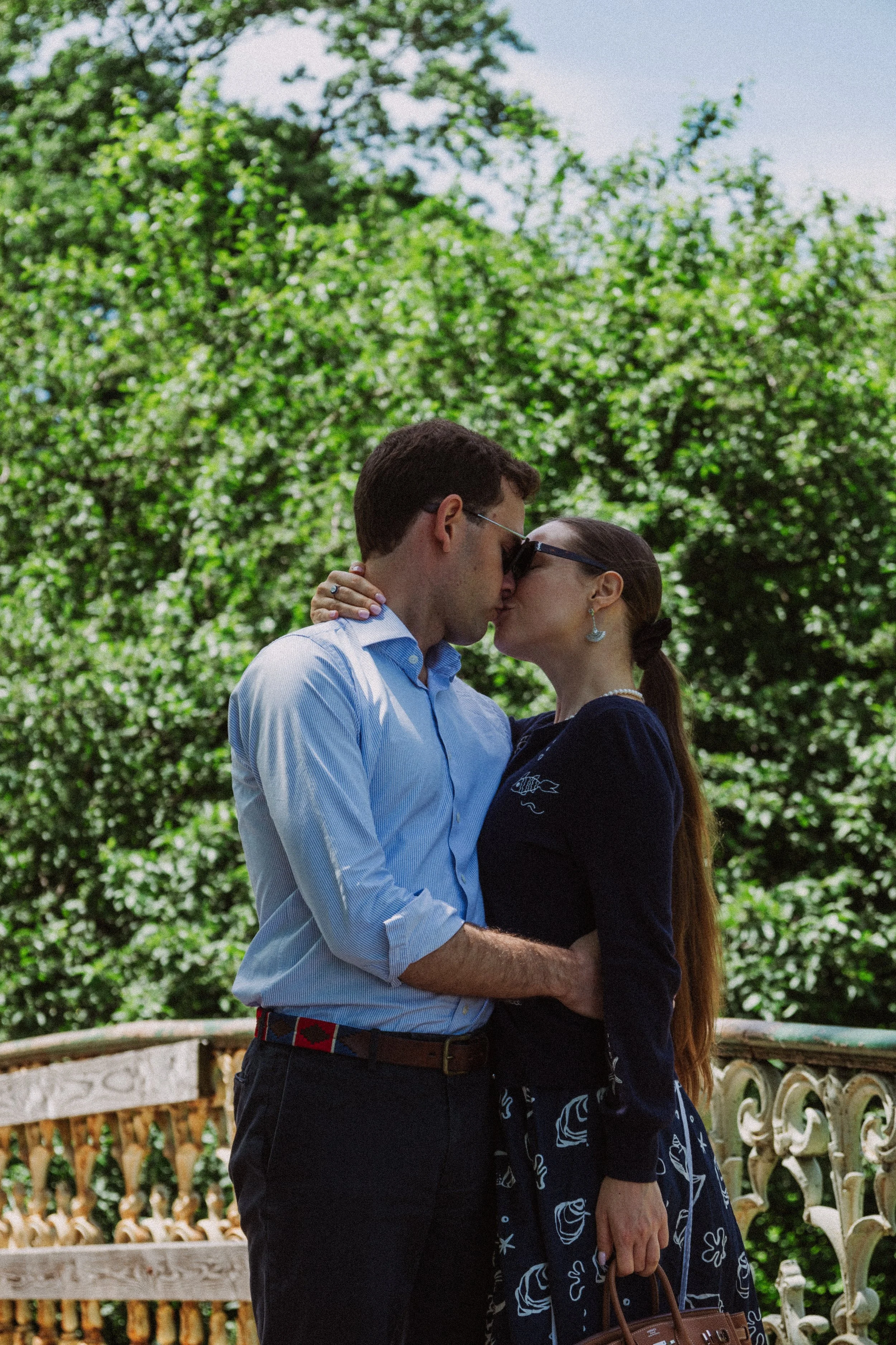 A man and woman embracing and kissing outdoors, with green foliage and a decorative white fence in the background.