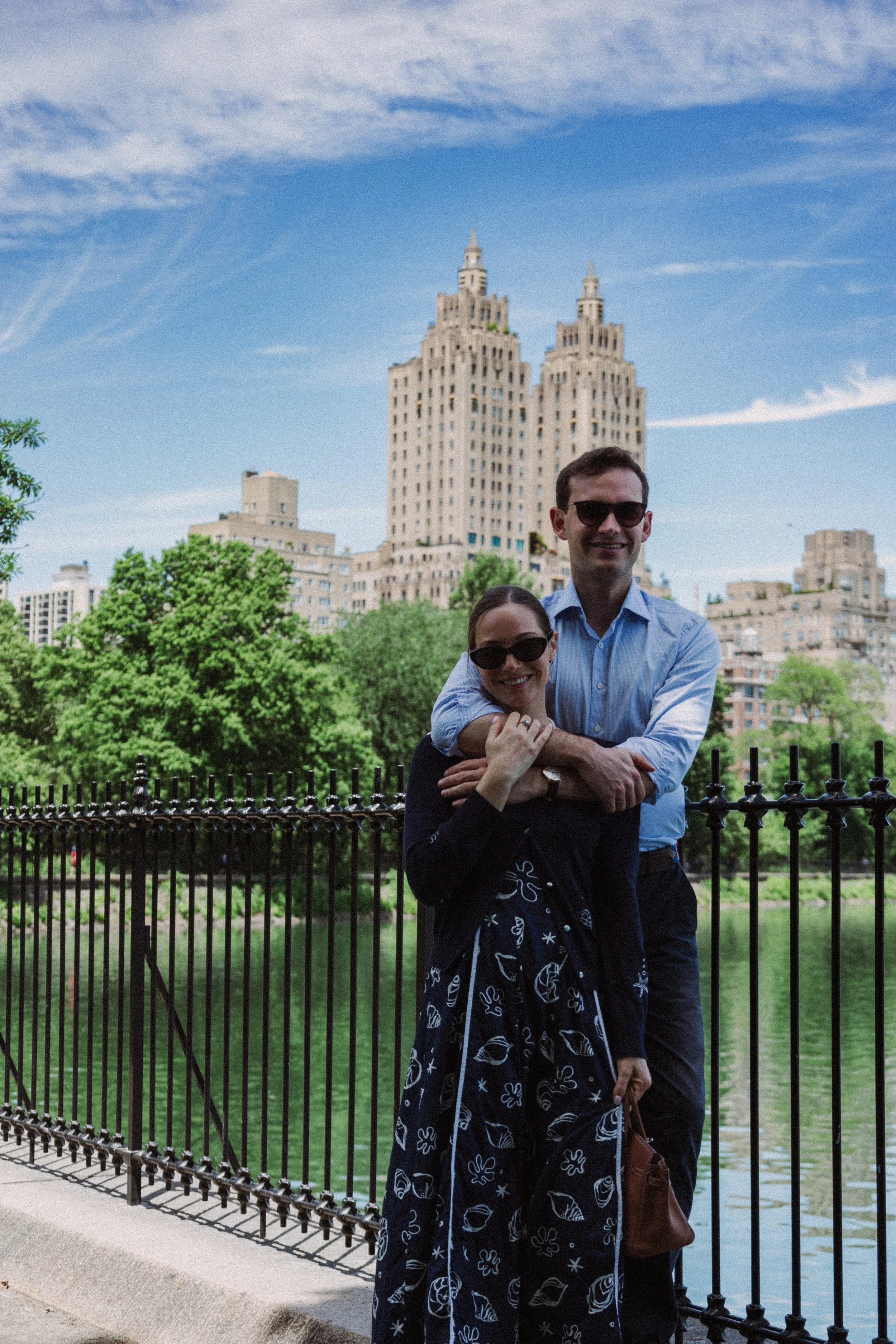 A man and a woman standing closely together outside by a fence, with a cityscape featuring two tall buildings and green trees in the background under a blue sky.