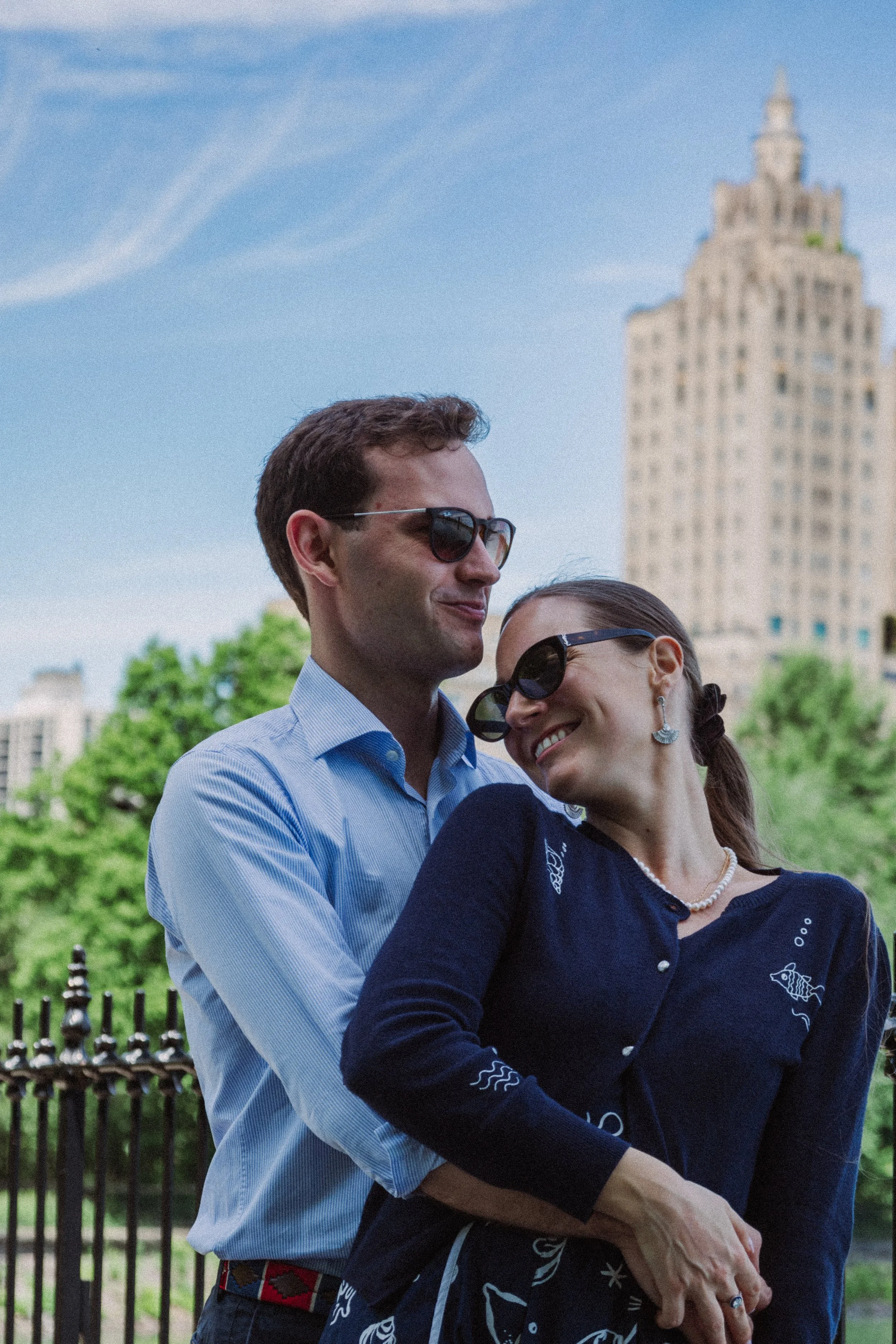 A man and a woman, both wearing sunglasses, are hugging in a park with a tall building in the background. The man is wearing a light blue shirt and the woman is wearing a dark blue sweater with embroidered designs.
