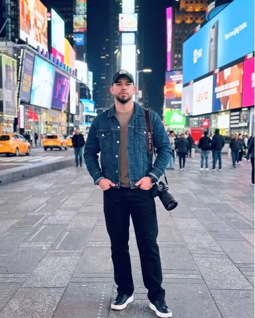 A man standing on a busy city street at night with bright digital billboards and yellow taxis in the background. He is wearing a black baseball cap, a denim jacket, and casual clothes, holding a camera.