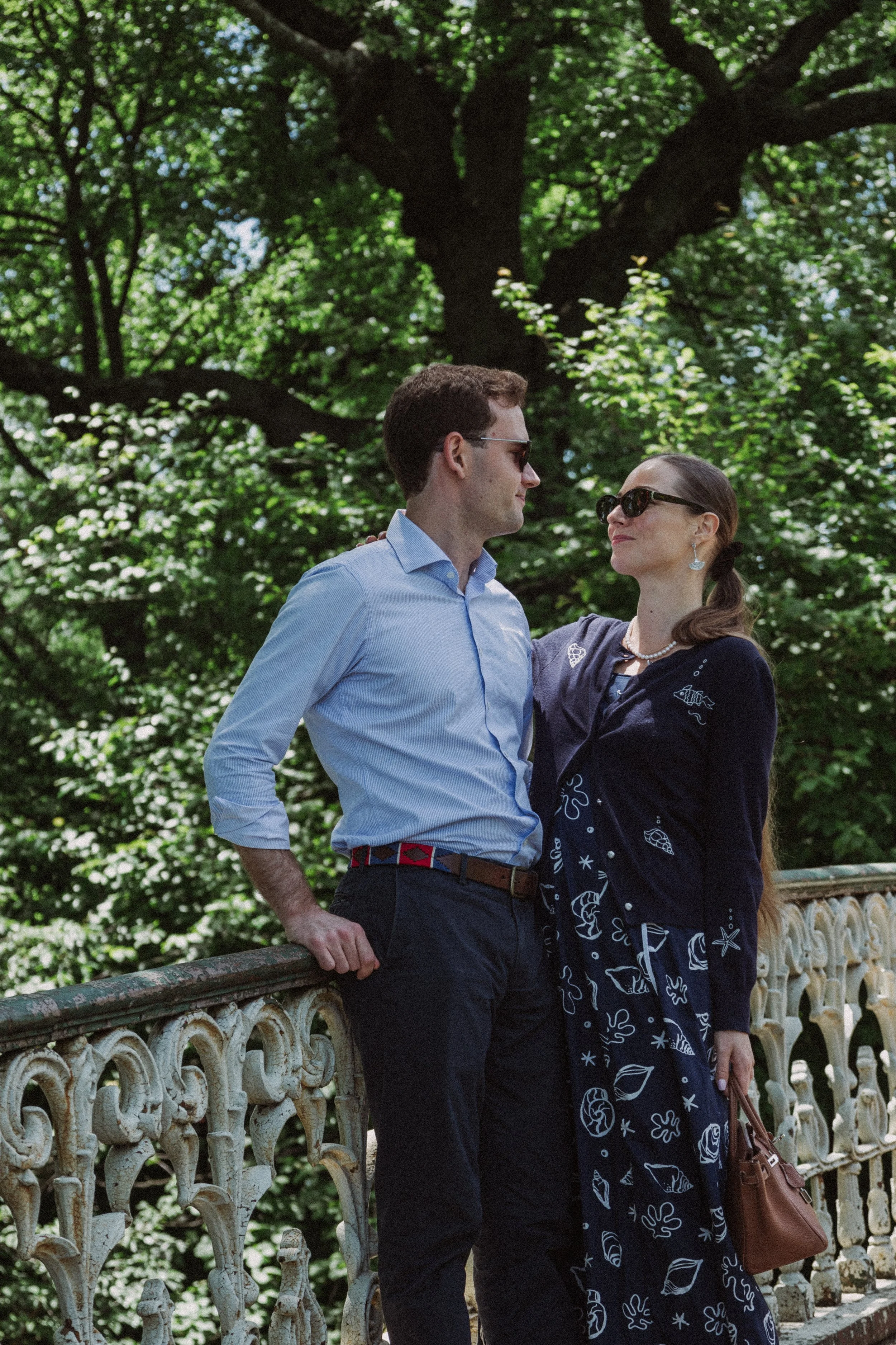 A man and woman standing close together on a decorated stone railing outdoors with green trees in the background, both wearing sunglasses and looking at each other.