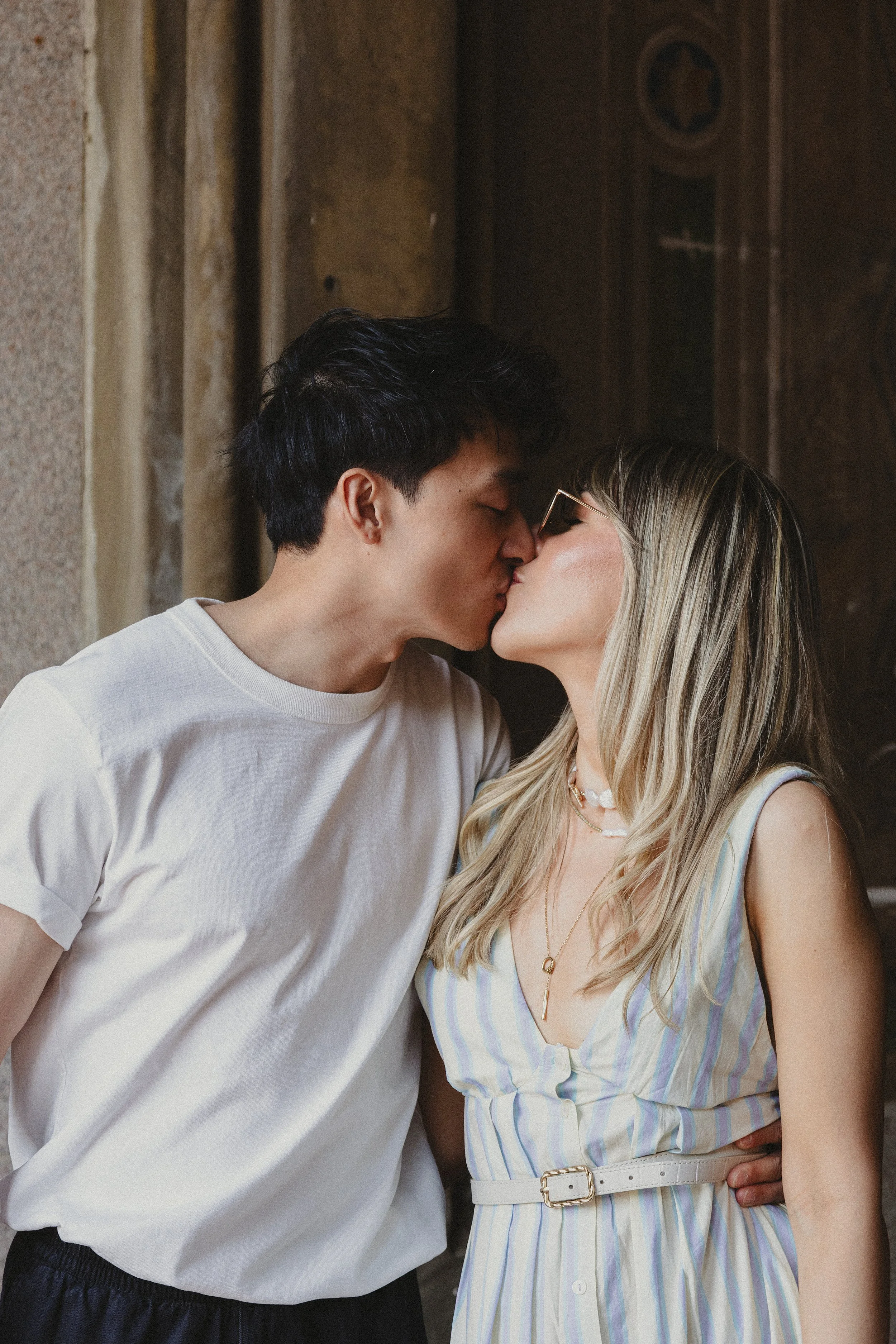 A man and woman are sharing a kiss indoors. The man has dark hair and is wearing a white T-shirt. The woman has long blonde hair with streaks, glasses, and is wearing a sleeveless striped dress with a belt. She has layered necklaces and is glancing u