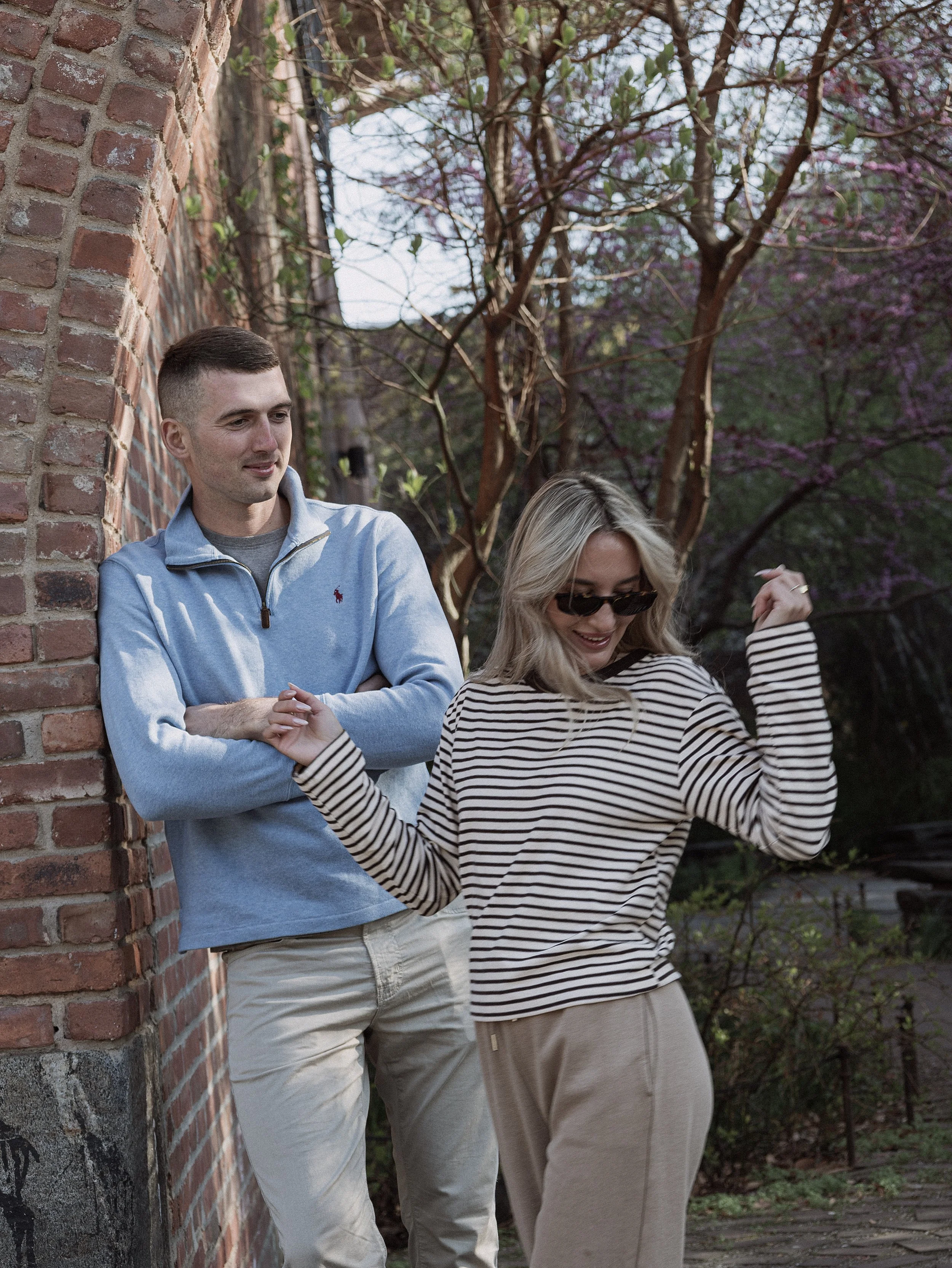 A young man and woman are standing outdoors near a brick wall and purple flowering trees. The man is leaning against the wall with his arms crossed, smiling slightly, wearing a light blue zip-up jacket and khaki pants. The woman, wearing sunglasses, 