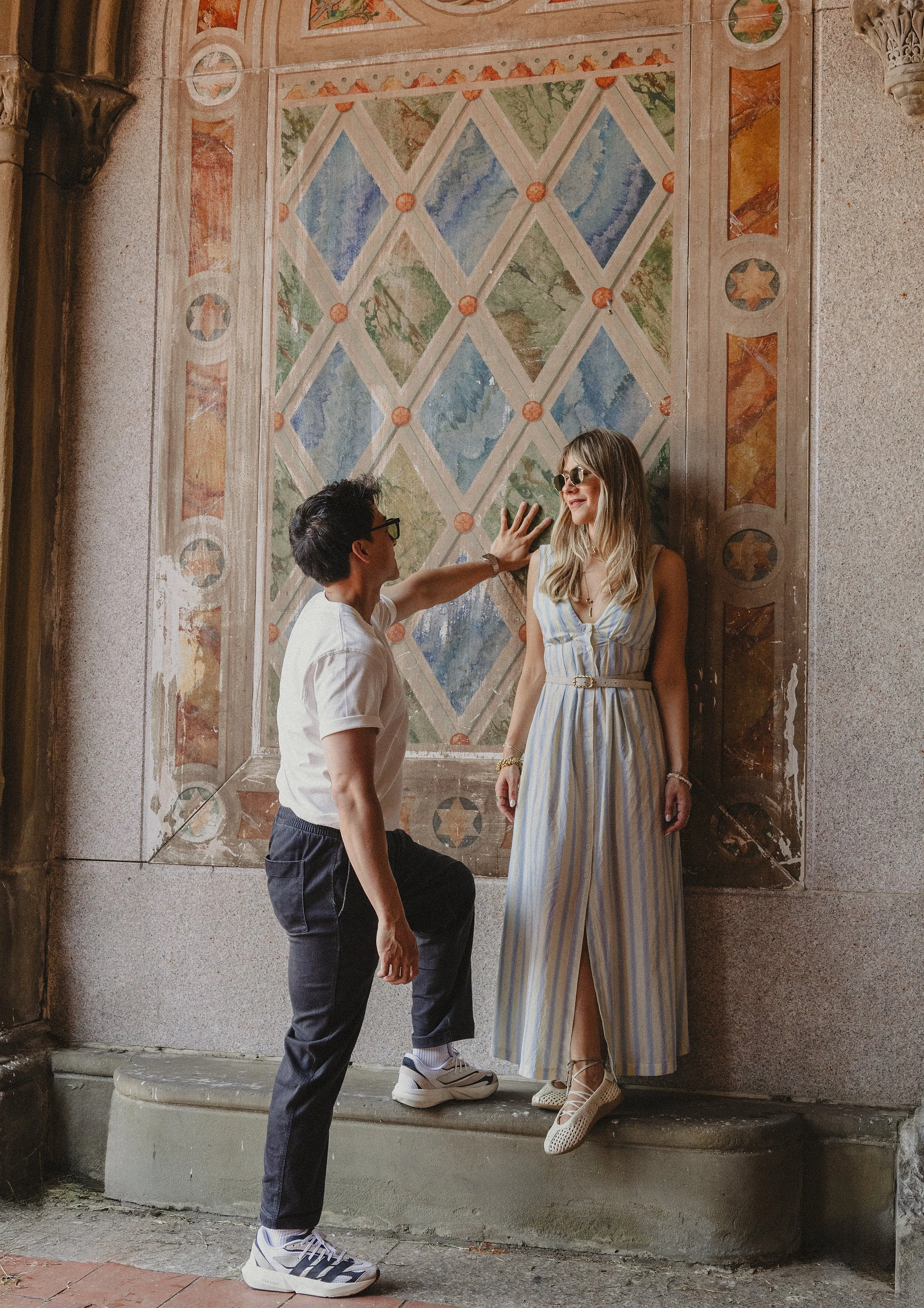 A man kneeling with one knee on a step and reaching out to touch a woman's cheek, who is standing with her back against a decorated wall. The woman is wearing a striped dress and sunglasses, and the man is wearing glasses and a white shirt. The setti