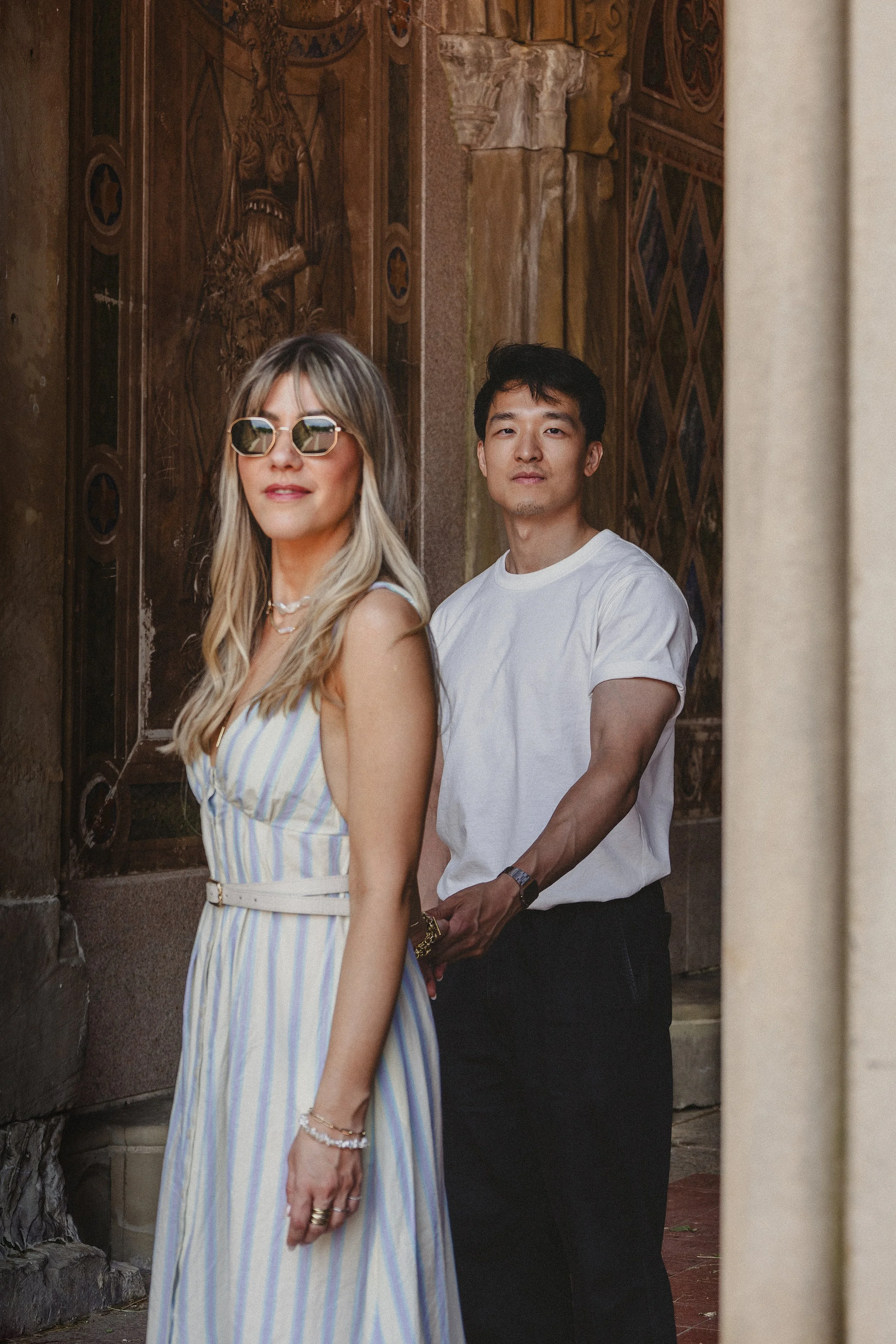 A woman with long blond hair wearing sunglasses and a striped dress stands in front of a man with black hair, wearing a white t-shirt, in an ornate building with painted wooden panels.