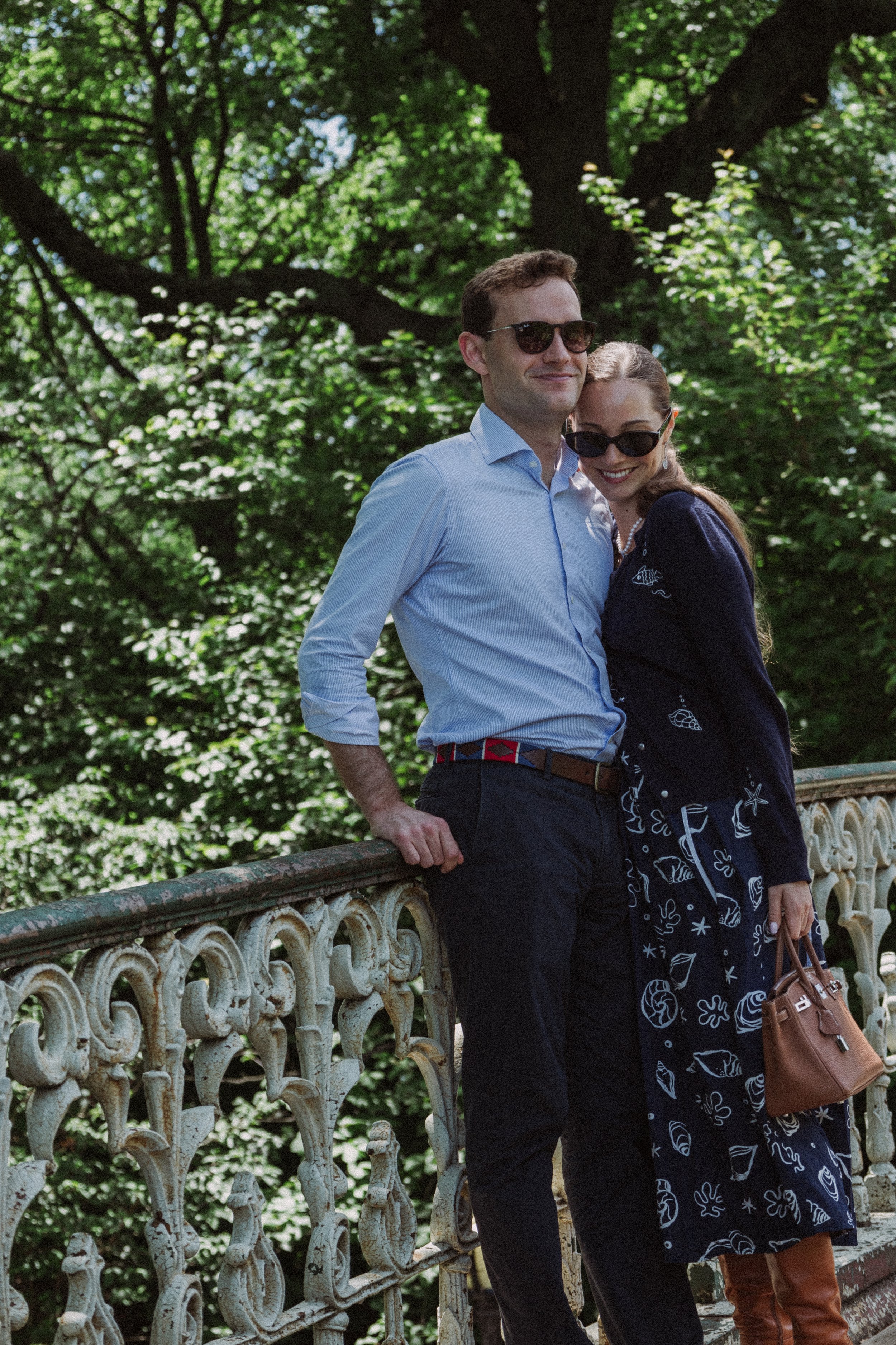 A man and a woman smiling and posing on a decorative stone railing in a lush green park, wearing sunglasses and casual clothing.