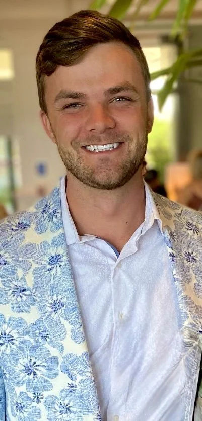 Smiling man with brown hair and a beard wearing a white shirt and a light blue floral blazer in a bright, indoor setting with green plants in the background.