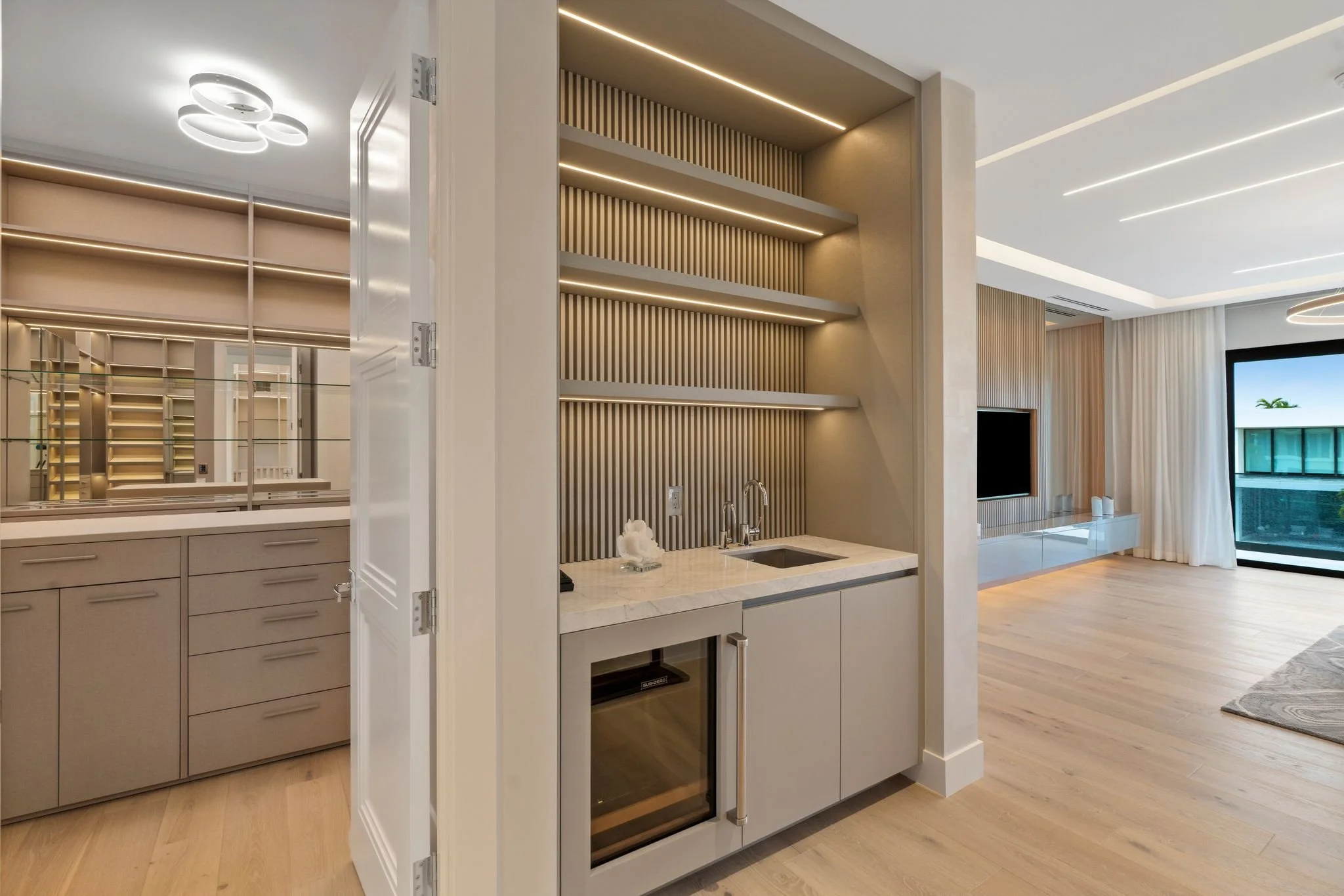 Modern interior with beige and white kitchen cabinetry, built-in oven, and a small sink, adjacent to an open living space with large windows and curtains.