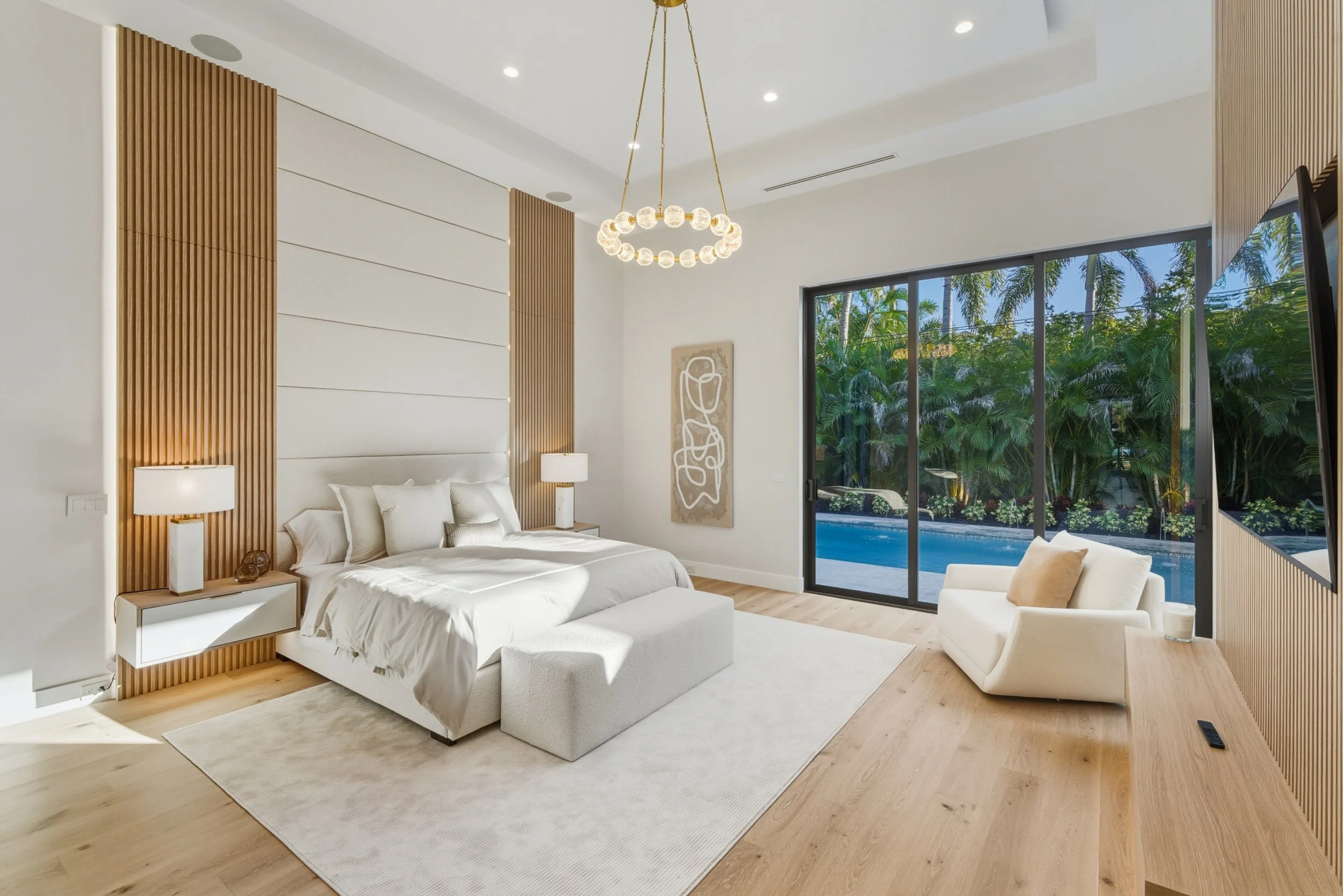 Modern bedroom with white bed, wooden slat wall accents, sliding glass door leading outside to lush green trees and pool, beige armchair, minimalistic decor, and a unique circular chandelier.
