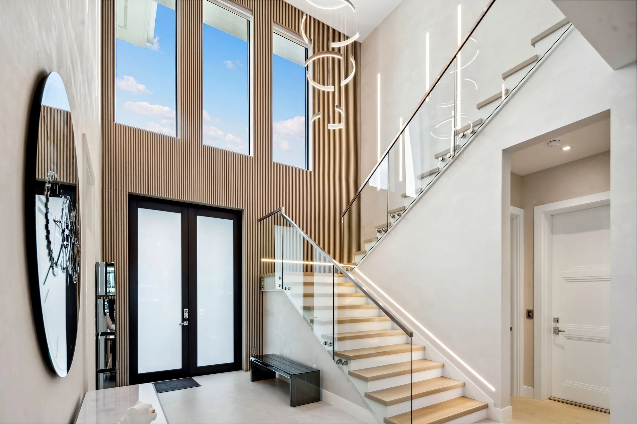 Interior of a modern house with a staircase, large windows showing a blue sky with clouds, a circular wall clock, and contemporary lighting fixtures.