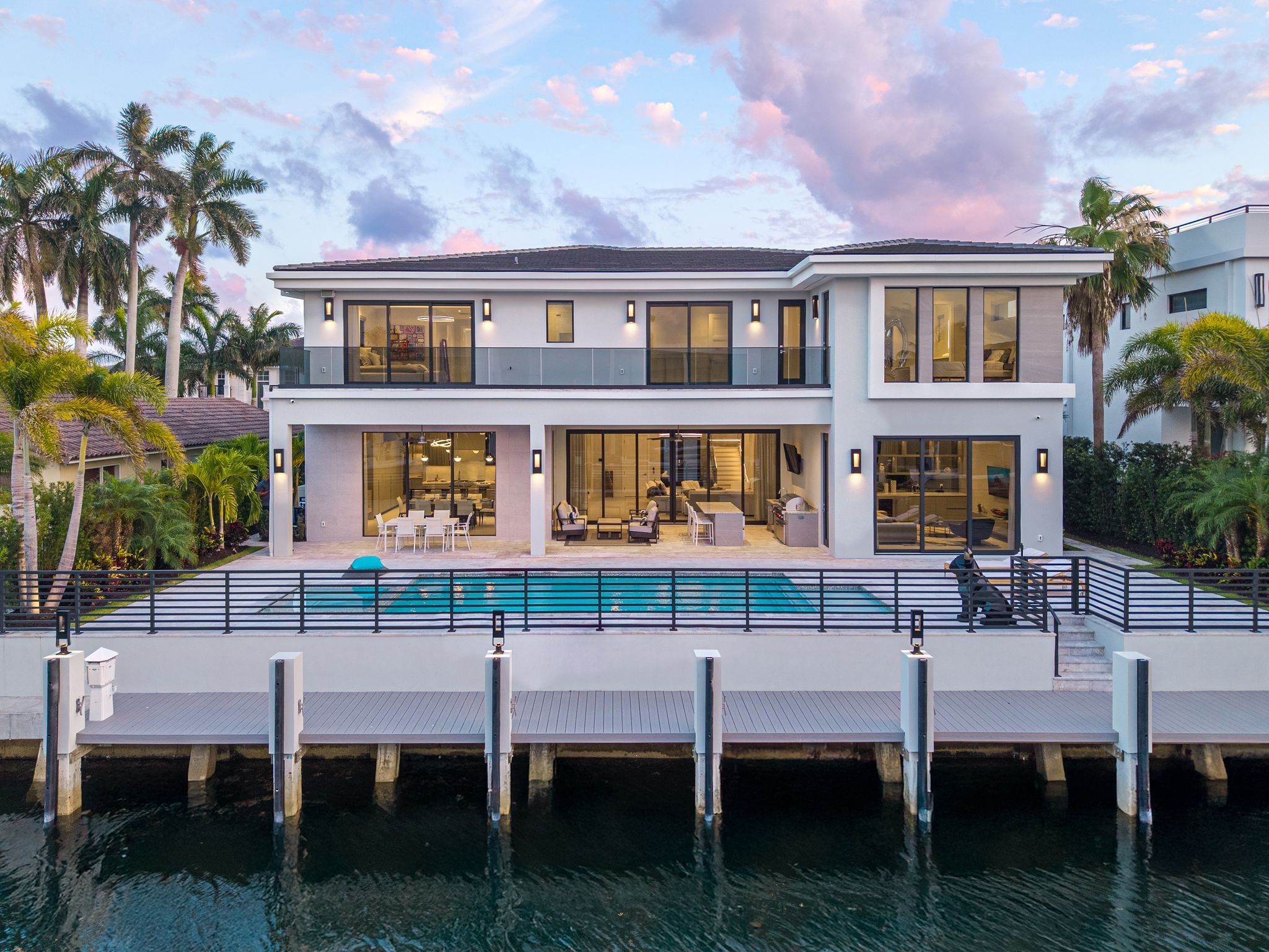 View of a modern two-story house with large glass windows and a balcony, situated beside a body of water with a dock in the foreground. The house is surrounded by palm trees and lush greenery, with a pink and blue sky overhead.