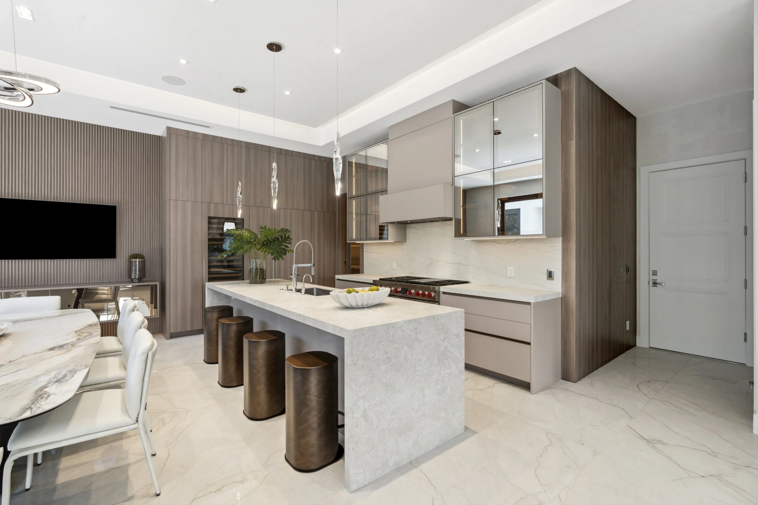 Modern kitchen with white marble island, beige cabinets, glass-fronted upper cabinets, and a wall-mounted television, featuring pendant lighting and decorative plants.